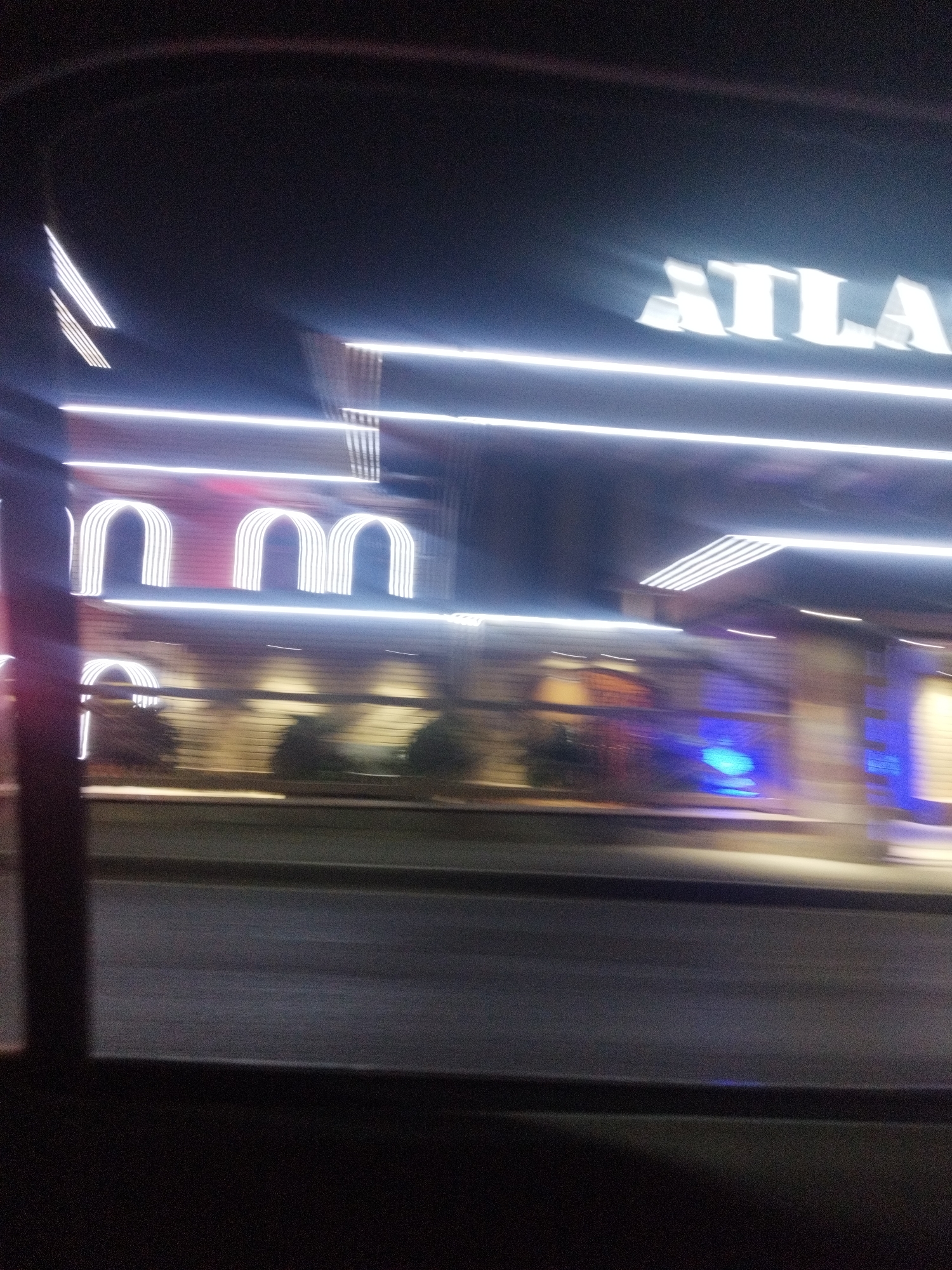This is a nighttime, motion-blurred photograph taken from inside a vehicle looking out at a building with bright neon lighting. The building has a sign with white letters that appear to spell "ATLANT" at the top. Below the sign are rows of illuminated arched structures, resembling doorways or windows, also outlined in white light. There are horizontal lines of white light running across the building's facade. In the lower right portion of the building, there are visible blue lights, possibly accent lighting or illuminated signage. The foreground shows a dark road surface with some faint lighting. The image is characterized by streaks of light due to the motion blur, suggesting the vehicle was moving at the time of the photograph. The location is identified as Shymkent, Kazakhstan.