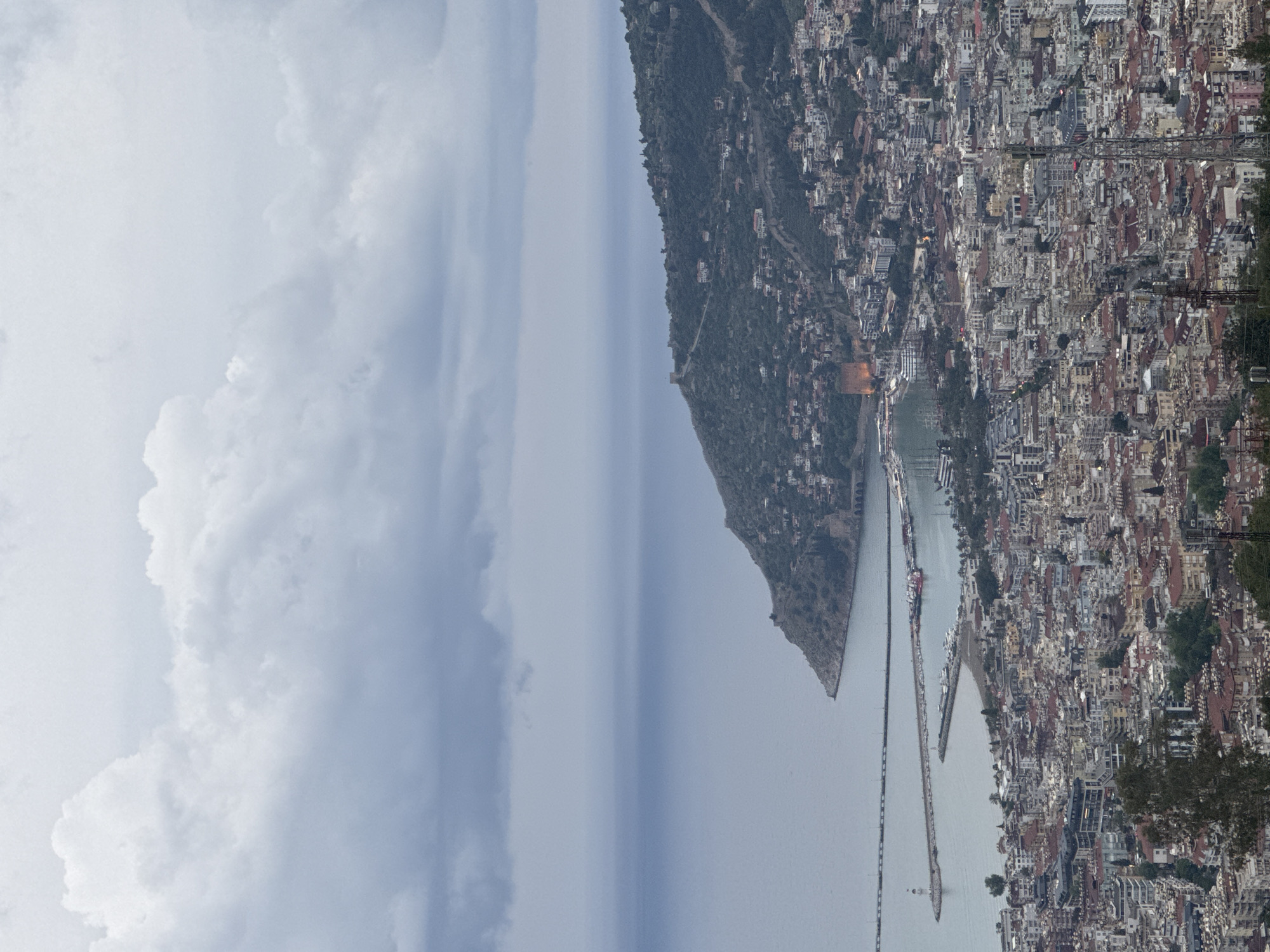 A high-angle, long-shot presents a coastal city of Unknown, Unknown. To the right, dense urban architecture dominates, with numerous buildings of varying heights and colors. A cable car system with utility poles is visible on the right edge of the image. In the center of the image, a peninsular promontory covered with green vegetation juts into the sea, separating the city and the sky. Two piers extend into the water, accommodating a mix of commercial ships. The sky is overcast, filled with varying shades of gray, suggesting an atmospheric or potentially rainy condition.