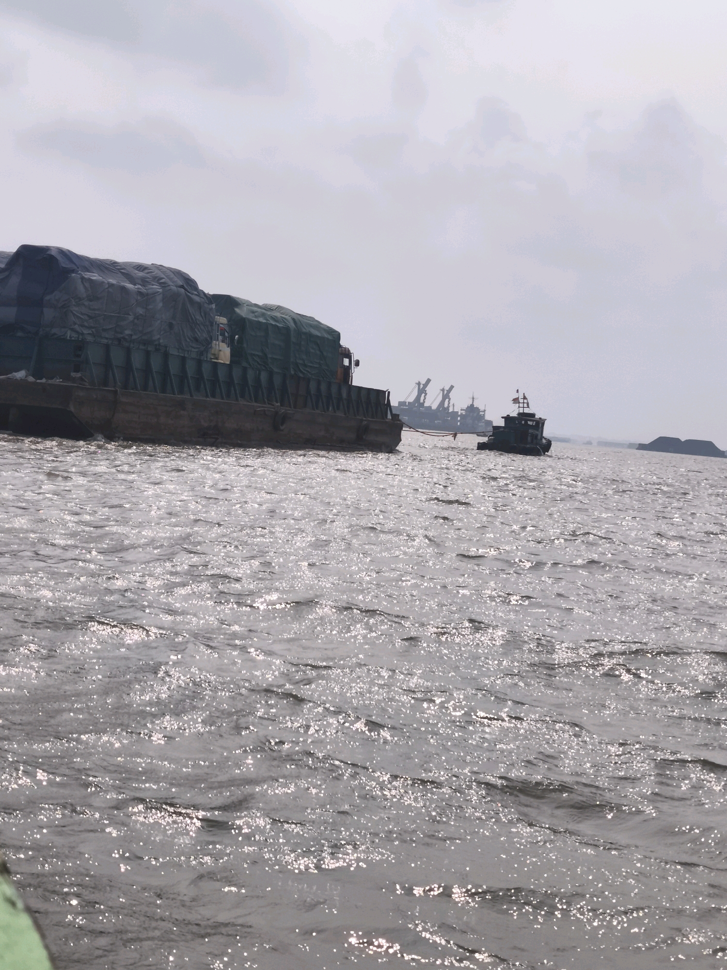 A wide shot depicts a body of water, likely a river, under a cloudy sky in Palembang, Indonesia. The water is choppy and reflects the overcast sky. In the foreground, a barge heavily laden with goods, covered by dark green tarpaulins, is moving from left to right. The barge appears to be towed or pushed by an unseen vessel, with a truck cab visible on its deck. Further in the distance, a smaller boat with an American flag is navigating the water. Behind this boat, the silhouettes of industrial cranes suggest a port or industrial area. A small, dark mound, possibly land or a constructed feature, is visible on the right horizon.