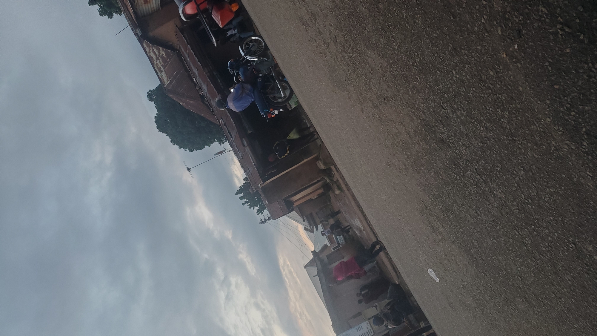 The image depicts a street scene in Jos, Nigeria, under an overcast sky. A paved road occupies the foreground, angled from the bottom right towards the top left. In the background, a row of buildings with corrugated metal roofs lines the street. Several people are visible, some standing and others seemingly in motion. On the road, a person is riding a motorcycle, wearing a blue shirt and a helmet. Another motorcycle, partially visible on the right, has a red attachment at the rear, suggesting it may be a commercial motorcycle (Okada). There are also people standing in front of the buildings and near the road. The overall impression is that of a typical urban environment in Jos, likely during a cloudy day.