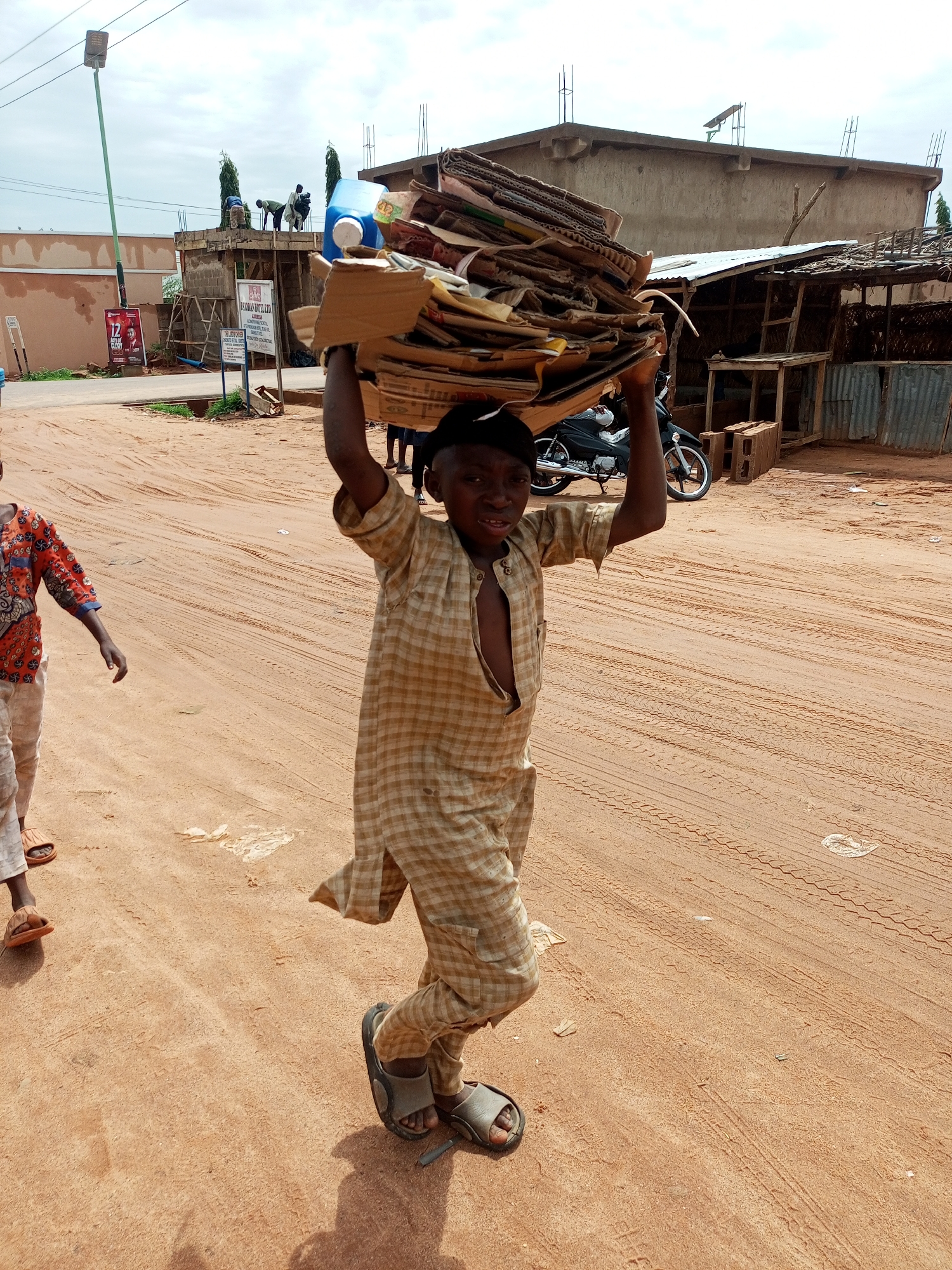 A young boy in a checkered tunic and trousers, wearing sandals, carries a large stack of cardboard on his head. He is walking on a dirt road in Tempure, Nigeria. Behind him, another child in a patterned shirt and trousers, also wearing sandals, walks in the same direction. In the background, construction is underway on a building. A motorcycle is parked near a small shop with a corrugated metal roof. The sky is overcast.