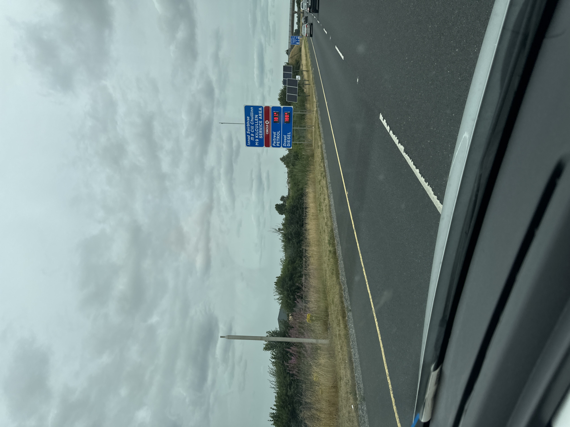 The image is a roadside view from inside a vehicle traveling on a highway in Ireland under a cloudy sky. A blue and red signpost is visible on the right side of the road, indicating "Ionad Seirbhise M9 CILL CHUILIUNN SERVICE AREA" (M9 Kilcullen Service Area). Below this, "CIRCLE K" is displayed in a red rectangle. Further down, prices for "Peitreal" (Petrol) are shown as 187.9 and for "Diesel" as 180.9. Several cars are visible on the highway in the distance. The area alongside the highway features dry grass, bushes, and a concrete pole on the left. There are also solar panels next to the signpost. The perspective suggests the photo was taken from the driver's or front passenger's seat.