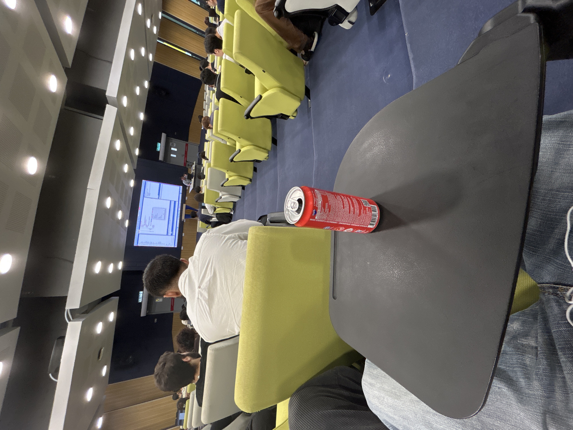 The image depicts the interior of a lecture hall, potentially in an educational institution in Kuala Lumpur, Malaysia. Numerous yellow chairs are aligned in rows, occupied by several individuals, predominantly students, facing towards a projection screen displaying data. A red soda can rests on a grey desk surface attached to one of the chairs in the foreground. The background reveals the hall's architecture, including artificial lighting.