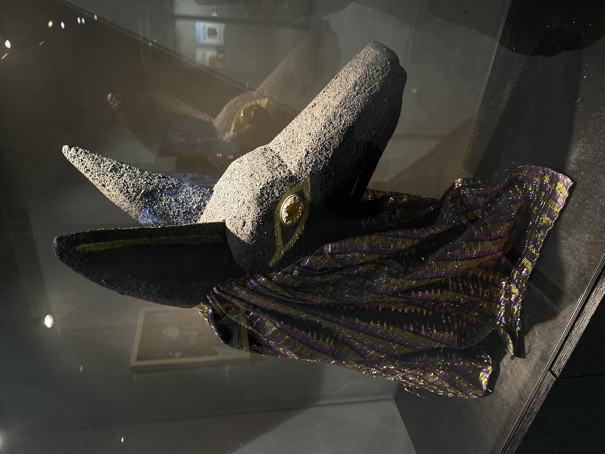 The image shows a close-up of an Anubis mask displayed in a glass case. The mask has a textured, dark gray surface and features large, pointed ears with a yellow stripe running up the center of each. The eyes are outlined in yellow, with a gold iris. The mask is draped with a shimmering fabric that has vertical stripes of purple and gold. The display case is dark, with reflections visible on the glass. In the background, out of focus, there appears to be artwork on a wall, possibly within a museum or gallery setting. The overall impression is of an ancient artifact or a theatrical prop.