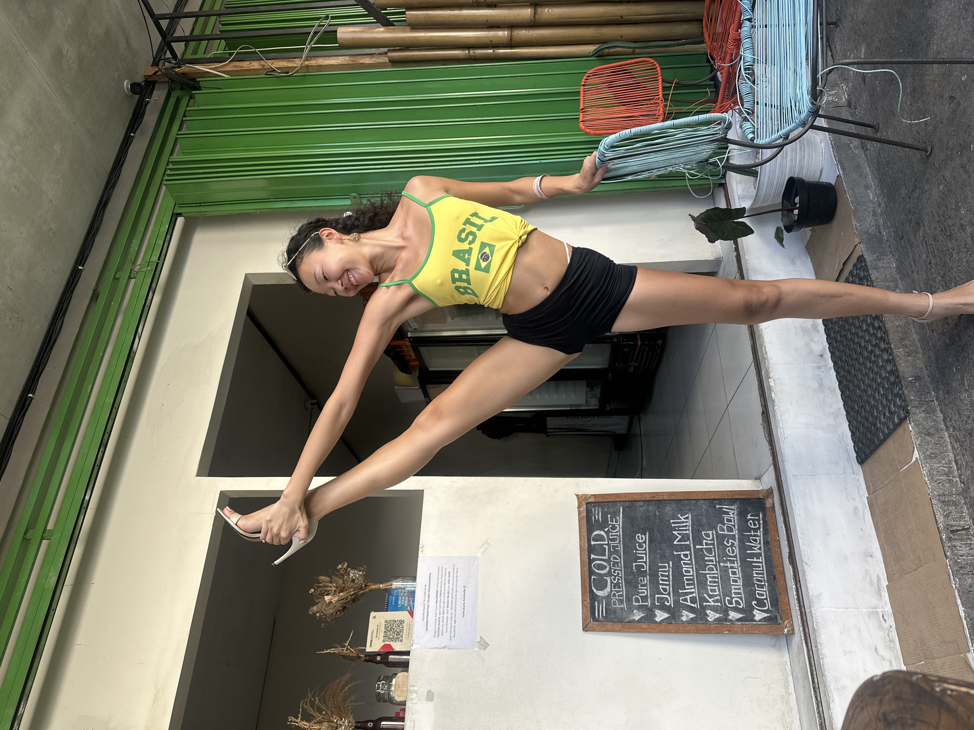 The image captures a woman in a yellow tank top with the word "BRASIL" printed on it and black shorts. She is executing a split between the entrance of a cafe, holding onto a blue chair on one side and resting her other leg on a ledge on the other side of the entrance. The cafe's storefront features a green corrugated metal awning, and a chalkboard menu listing "COLD PRESSED JUICE," "Pure Juice," "Jamu," "Almond Milk," "Kambucha," "Smooties Bowl," and "Coconut Water." The location is in the city of North Kuta, Indonesia.
