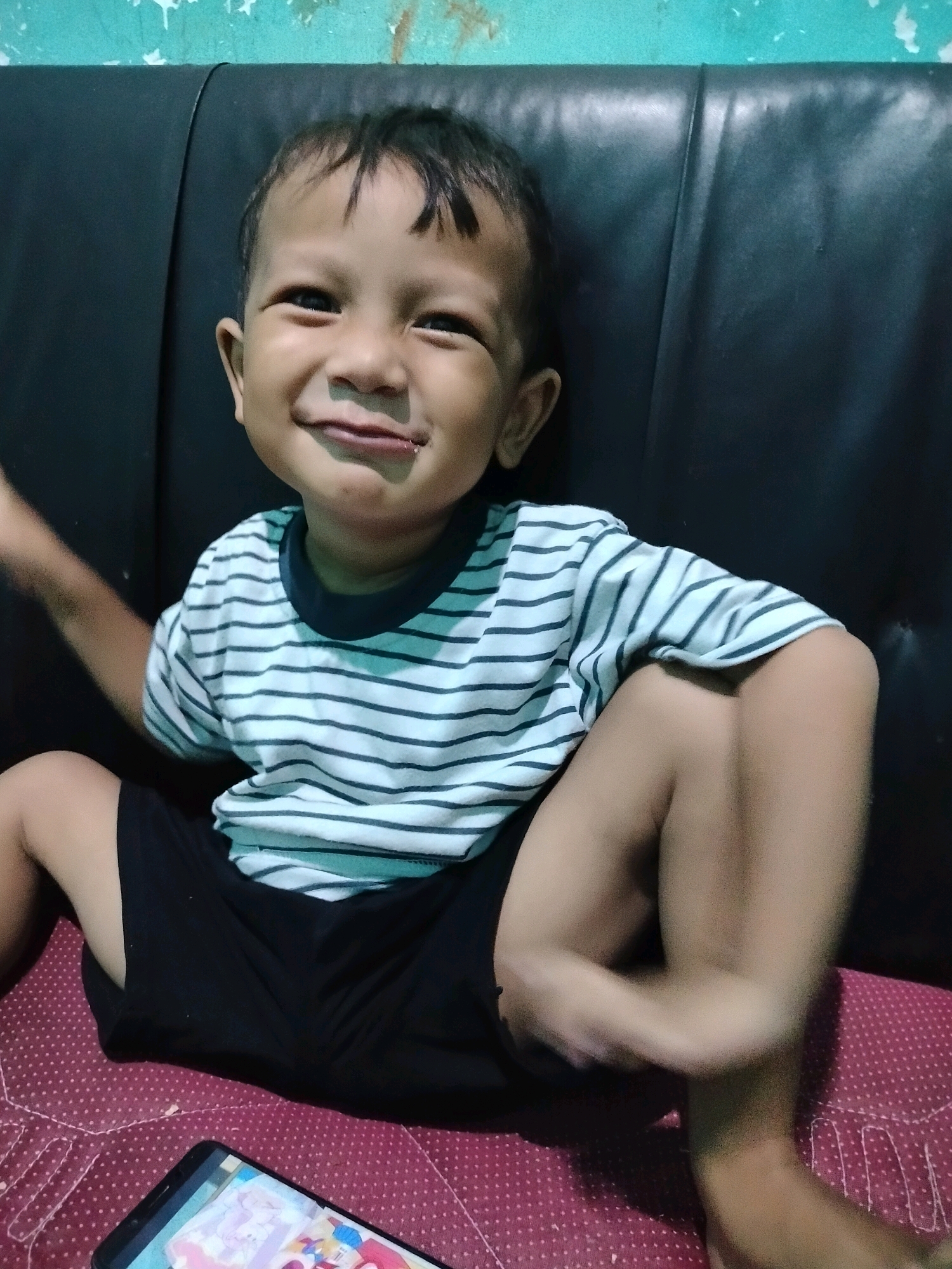 A young East Asian boy, estimated to be between 3-5 years old, is pictured indoors, seated with his legs crossed. He is wearing a striped t-shirt with horizontal dark and light blue stripes, and black shorts. The boy is looking towards the camera with a wide, joyful smile, his eyes crinkled at the corners. A small amount of food residue is visible on his lower lip. He is positioned in front of a dark, possibly leather, cushioned surface, which appears to be part of a sofa or bench. Behind this surface, a teal-colored wall with some white paint drips is visible. In the lower left of the frame, a smartphone is lying on a red surface with a subtle white polka dot pattern. The lighting suggests an indoor setting, likely in the evening or with artificial light. The scene appears to be a casual, domestic environment.