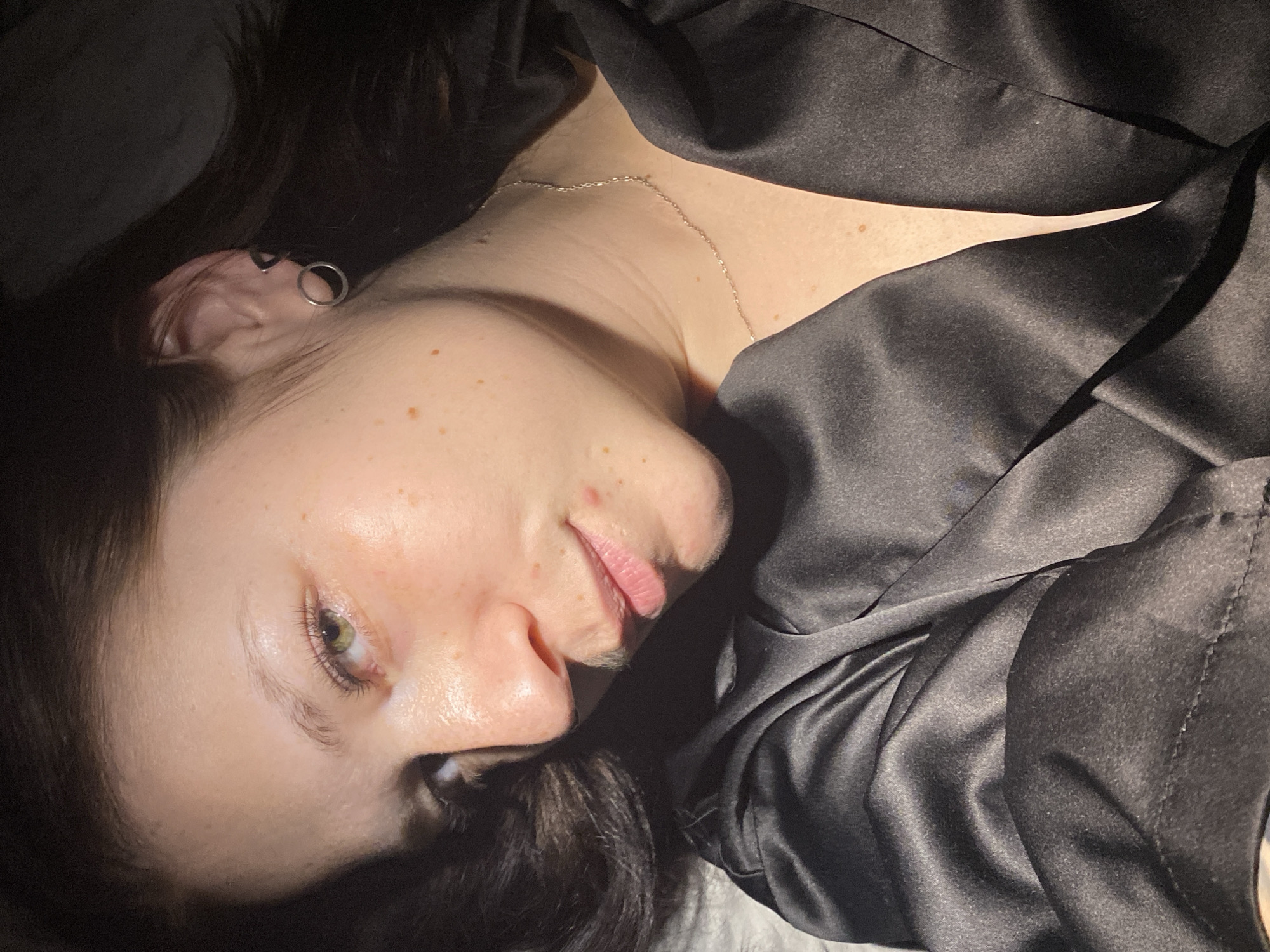 A person with dark hair and light skin lies down, facing upwards and to the right of the frame. They are wearing a black silky garment. A delicate silver chain necklace is visible on their neck. The person has multiple piercings in their left ear. The environment appears to be dimly lit. There is no information to suggest that this scene is in Chaikovsky, Russia.