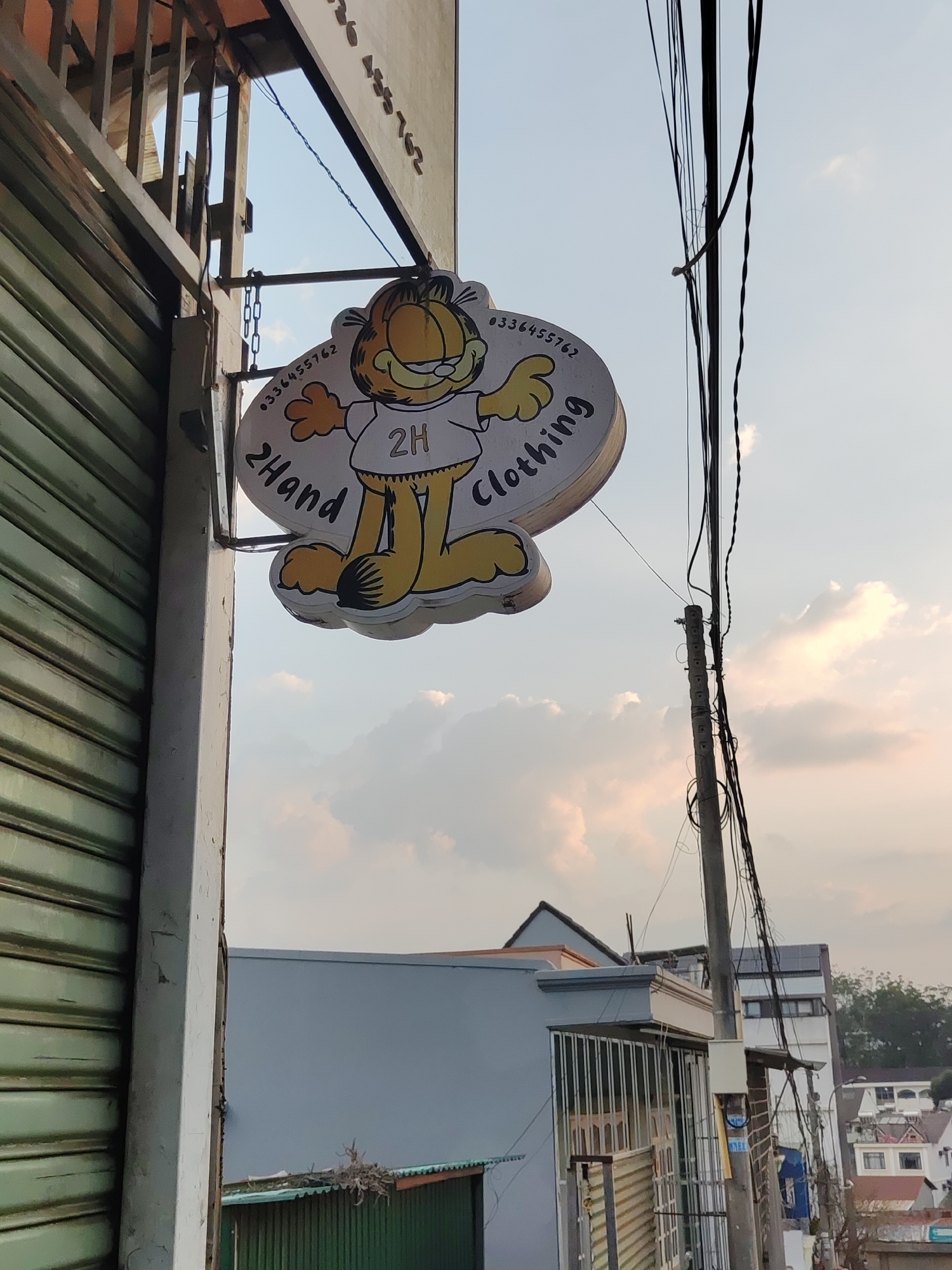 An outdoor, medium-shot photo depicts a storefront in Da Lat, Vietnam, featuring a sign for "2Hand Clothing" with a cartoon Garfield character. The sign is circular with Garfield wearing a t-shirt labeled "2H". To the left is a closed, green, corrugated metal roll-up door and a weathered, white doorframe. Overhead, the top of the building is partly visible with corrugated metal. In the background are buildings and a utility pole with multiple wires. The sky is overcast with pale orange and white hues.