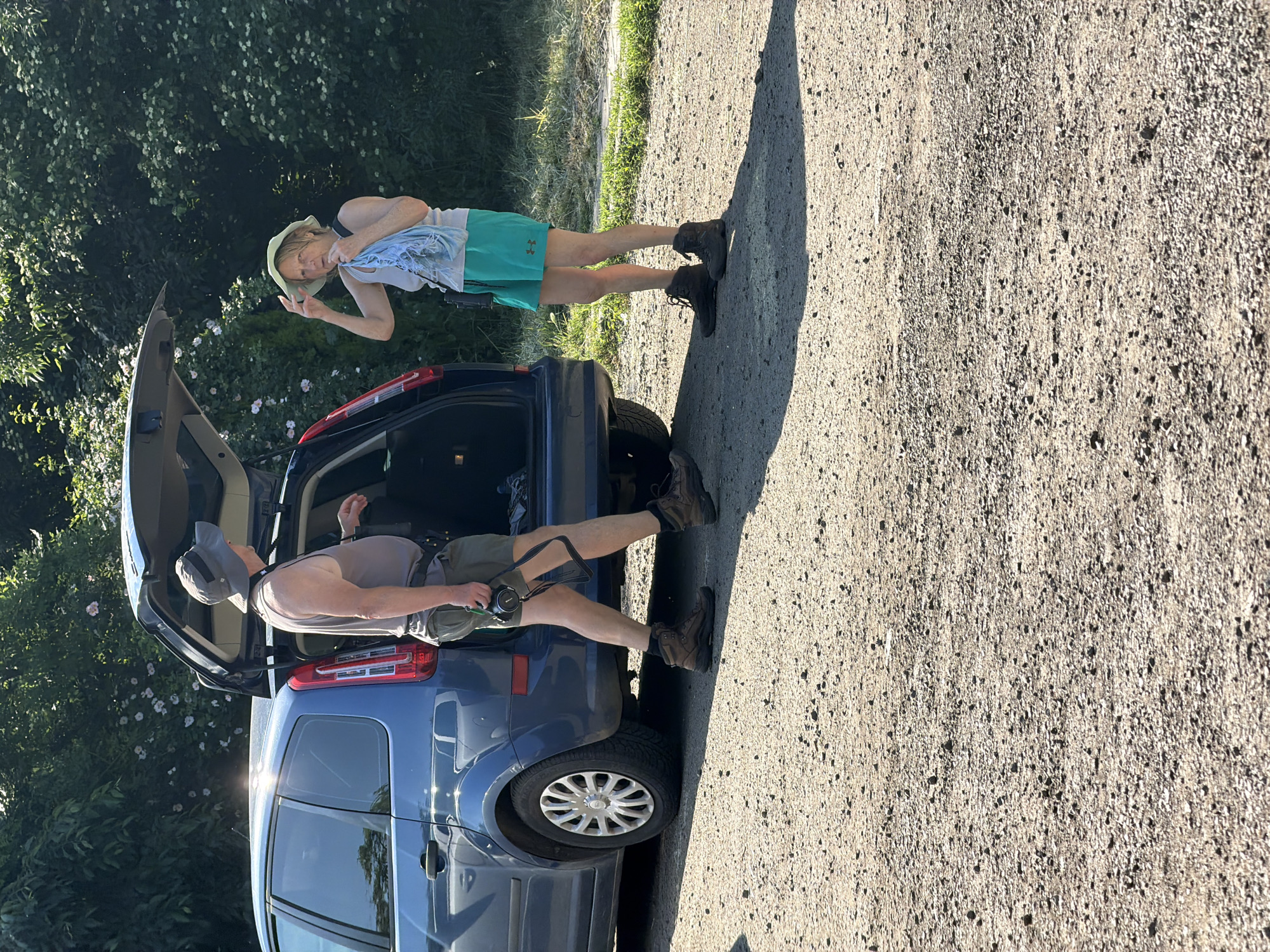 Two individuals are positioned near a blue hatchback car, likely a Ford Focus, parked on a gravel surface. The car's rear hatch is open. One person is seated inside the trunk. The second person is standing near the passenger side rear. Both appear to be dressed for outdoor activity, with hats, shorts, and boots. The background includes trees and shrubs, suggestive of a rural or semi-rural location. There is a bright sunny, with long shadows in the scene. The precise location in Burgas, Bulgaria cannot be ascertained from this single image.