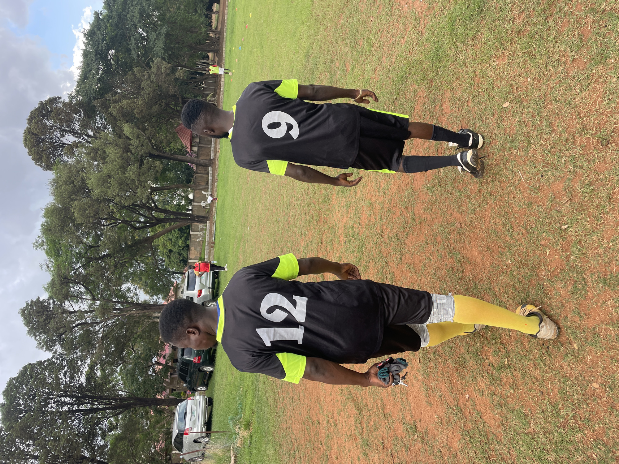 The image captures two men standing on an outdoor sports field, viewed from behind. Both are dressed in black sports jerseys with neon yellow accents on the sleeves and sides, paired with black shorts. Their attire suggests they are involved in athletic activity, likely soccer or football.
The man in the foreground wears the number "12" prominently displayed in white on the back of his jersey. He has bright yellow socks covering his calves and light-colored athletic shoes. In his left hand, he holds a small collection of items, including what appears to be keys and a dark-colored cloth or glove.
Further into the frame, the second man wears jersey number "9" in white. He has black socks and black and white athletic shoes. Both individuals are standing on a ground surface that is a blend of green grass and reddish-brown earth, typical of some outdoor playing fields.
The background features a dense line of tall, mature green trees under a partly cloudy sky. Beneath the trees or slightly beyond them, several cars are parked, indicating accessibility to the field. In the distance, other figures in sports attire are faintly visible, suggesting more people are present or engaged in activities on the field. The overall lighting indicates it is daytime, possibly under overcast conditions or diffused sunlight. This scene likely takes place in Jos, Nigeria.