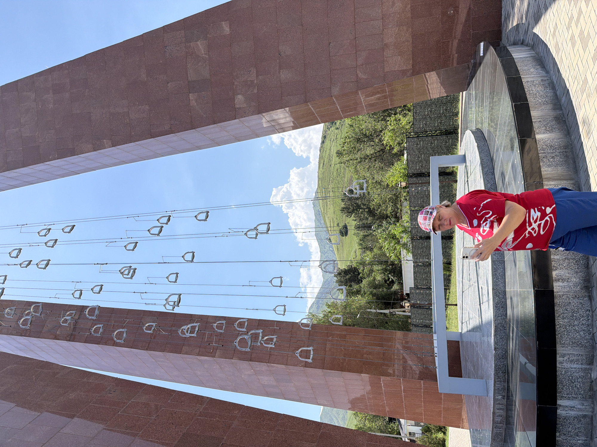 A woman stands in front of a monument. The monument is comprised of two tall, reddish-brown structures that lean inwards, with numerous stirrups suspended by wires from a central point. The woman is wearing a red shirt with white abstract designs, blue pants, and a patterned baseball cap. She is holding a smartphone in her hands and looking down at it. The background shows a bright blue sky with some clouds, green rolling hills, and trees. The ground in the foreground is paved with bricks, and there is a circular base to the monument with reflective surfaces.