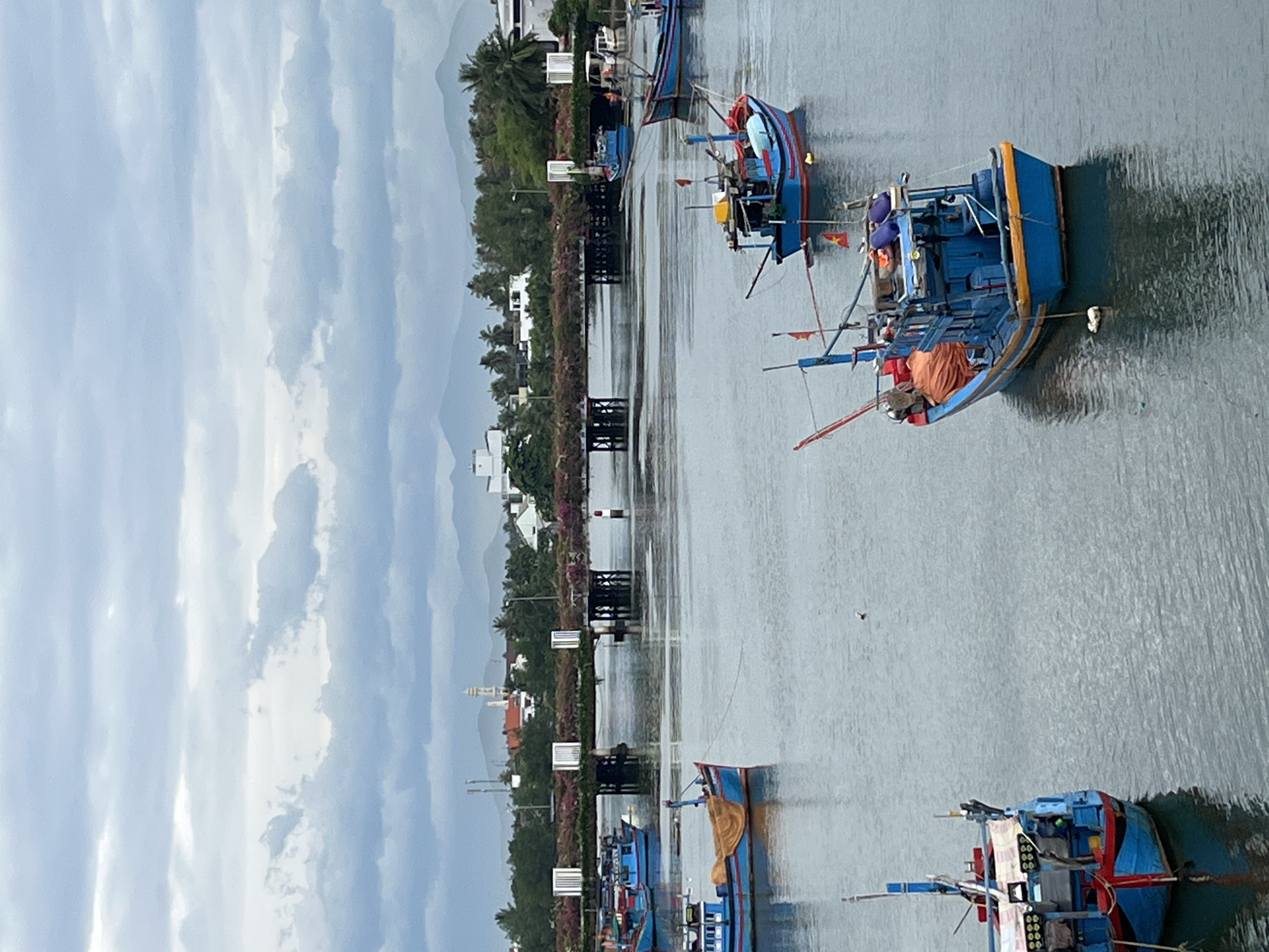 The image captures a waterway in Nha Trang, Vietnam, with several blue fishing boats docked or anchored. The sky is overcast with heavy clouds, casting a diffused light over the scene. In the background, a bridge spans the water, lined with greenery and structures, suggesting an urban or semi-urban setting. Distant mountains form a hazy backdrop. The water's surface is calm, reflecting the sky and the boats. One boat in the foreground is particularly detailed, showing its blue hull, orange netting, and various equipment. Another boat to the left, also blue, is partially visible. Several other boats are scattered across the waterway, varying in size and visibility. The overall impression is one of a working harbor or a scenic river view within the city.