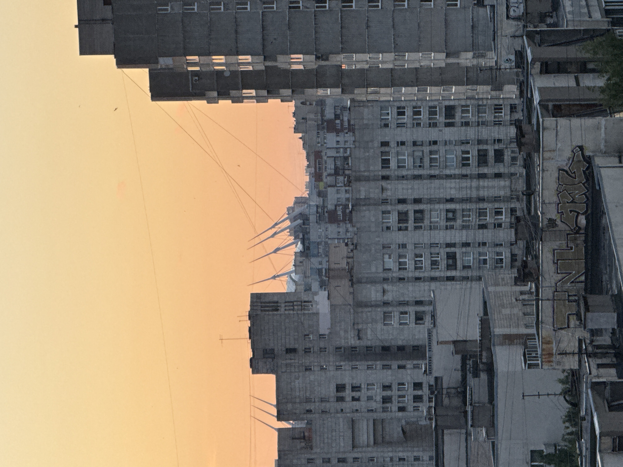 The image depicts a cityscape of Saint Petersburg, Russia, during what appears to be sunset or sunrise. The sky is a gradient of warm orange and yellow hues. In the foreground and midground, there are multiple multi-story residential buildings, characterized by their grey, concrete facades and numerous windows. Some of these buildings have a repetitive, brutalist architectural style.
In the background, the distinctive white, tent-like structures of the Gazprom Arena (also known as the Zenit Arena) are visible. The arena is a prominent landmark located on Krestovsky Island in Saint Petersburg. The presence of these structures confirms the location.
The roofs of some buildings in the foreground display various rooftop structures, including ventilation shafts, antennae, and some graffiti. There are also power lines crisscrossing the sky. The overall impression is of a densely populated urban environment with a mix of residential blocks and modern sporting infrastructure. There are no people visible in the image. The scene is static, with no discernible actions or interactions taking place.