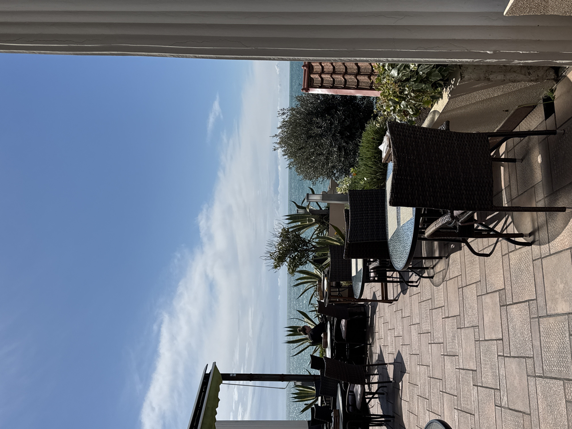 The image shows an outdoor cafe terrace with wicker chairs and small round tables. The terrace is tiled in a rectangular pattern. The cafe overlooks the sea and sky, with some trees and greenery interspersed along the edge. The sky is blue with some scattered clouds. There is a glimpse of a building with a red-tiled roof. A person is seated at one of the tables. It can be assumed that the location is in a coastal area in the city of Sochi, Russia.