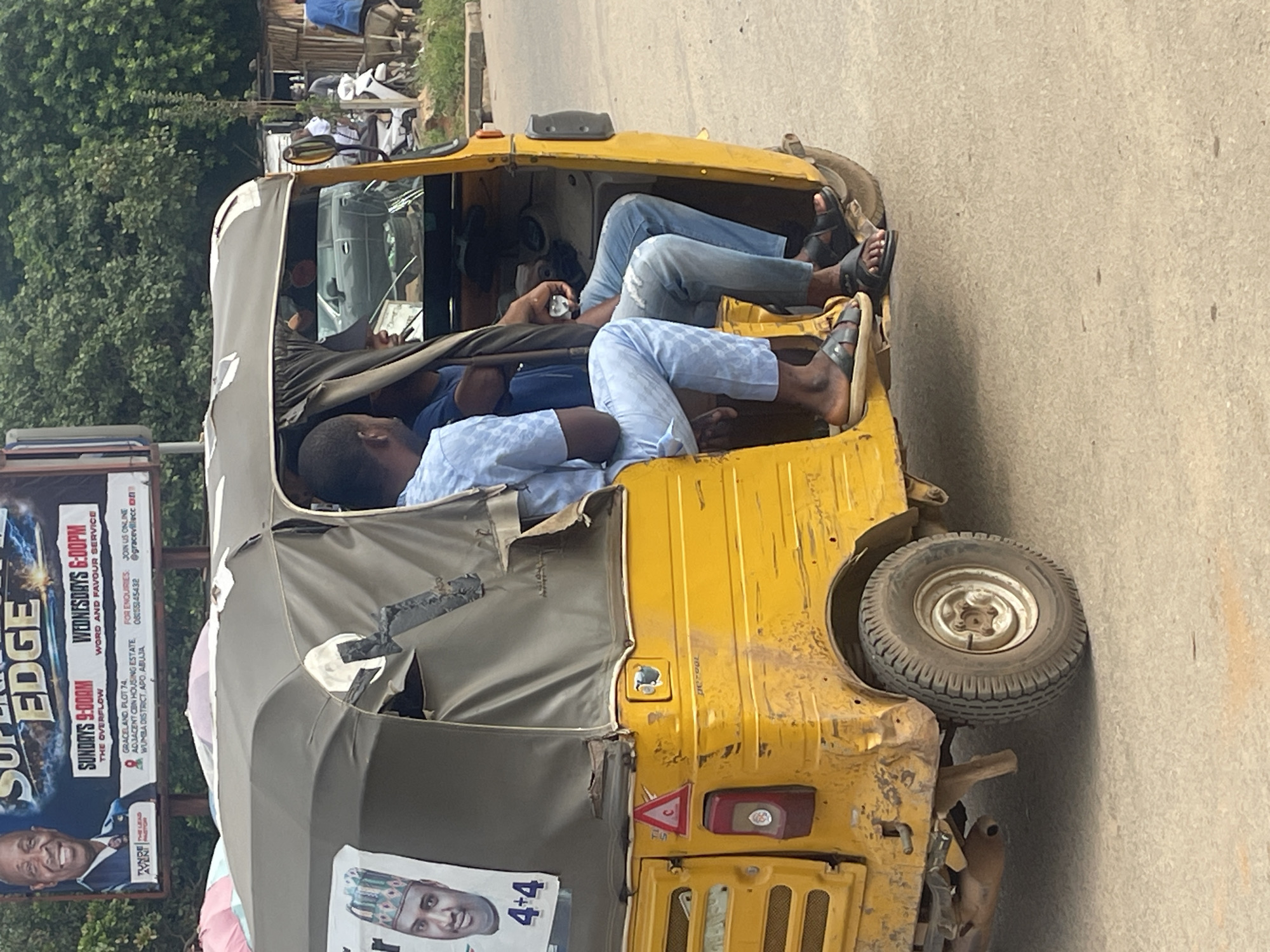 In Abuja, Nigeria, a yellow auto-rickshaw, commonly known as a "Keke," is seen from the side. The Keke is carrying three passengers: a driver and two passengers seated behind him. The driver, wearing a light blue shirt and ripped jeans, is operating the vehicle. The passenger directly behind him is wearing a blue shirt, and the passenger further back is partially obscured but appears to be wearing a blue outfit. The exterior of the Keke shows signs of wear and tear, with some rust and dents. A poster is visible on the side of the Keke, featuring a man's face and the text "4+4." The scene appears to be on a street with a paved surface. In the background, there are trees and a billboard advertising a church service, indicating this is likely a commercial or residential area of Abuja.