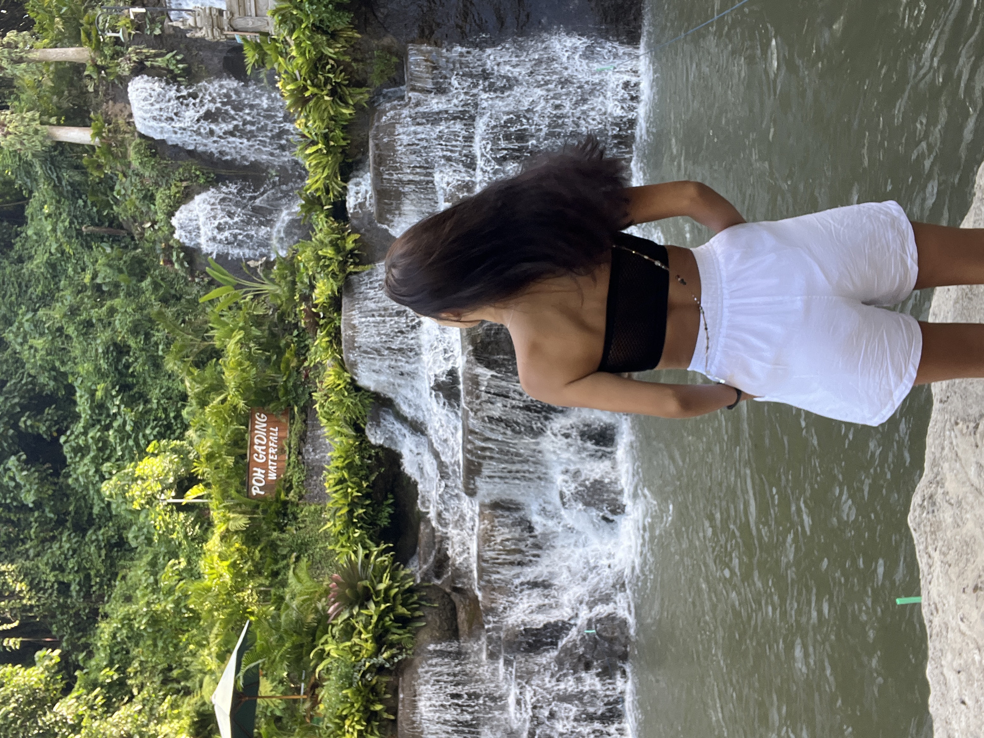 A vertical, medium shot captures a woman standing in front of Poh Gading Waterfall in Sukawati, Indonesia. She faces away from the camera, her dark hair cascading down her back. She wears a black top and white shorts. The waterfall cascades down a rocky, plant-covered terrain into a body of water. Lush greenery surrounds the waterfall, and a sign reading "POH GADING WATERFALL" is visible. The water appears turbid. The woman seems to be standing on a stone edge.