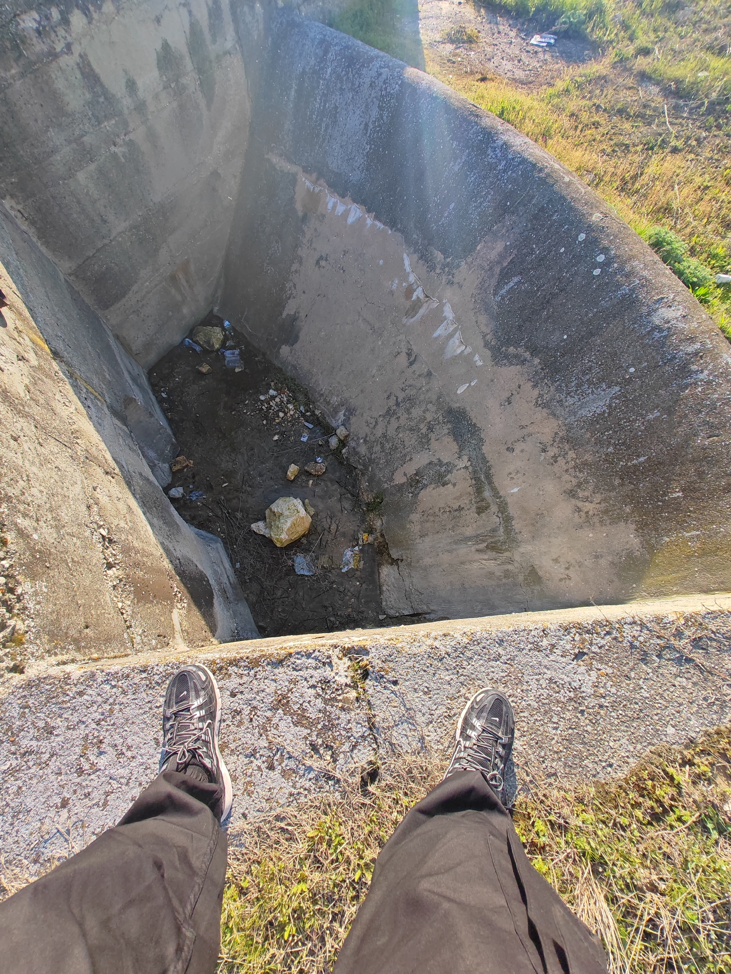 The image, taken from an elevated perspective, shows a person standing at the edge of a concrete structure in Shumen, Bulgaria, looking down into a deep, possibly abandoned or disused, concrete drain. The person is wearing dark pants and athletic shoes. Below, the drain contains water, mud, rocks, and some litter. The drain's concrete walls are stained and show signs of water damage. Outside the structure, the ground is covered in grass and some weeds, indicating an outdoor environment.