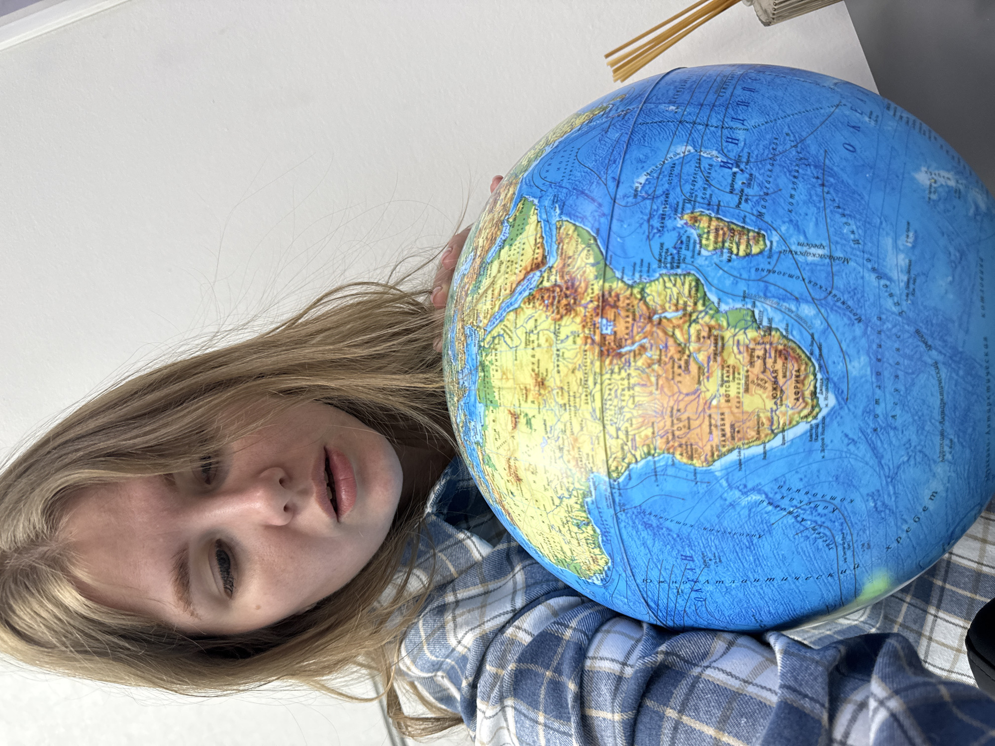 A person with long, light brown hair is holding a globe in front of them. The globe depicts Africa and the surrounding oceans. The person is wearing a blue and white plaid shirt. In the background, there's a white wall, some straws, and a piece of paper. The location is unknown.
