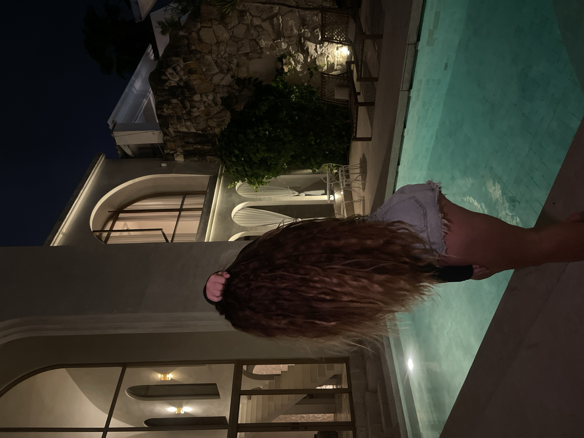 A person with long, curly, reddish-brown hair is hunched over, touching their hair with their right hand. They are wearing a black long-sleeved shirt and light denim shorts. The person is positioned at the edge of a turquoise swimming pool, with the pool occupying the lower right portion of the frame.
The background features a modern villa with a grey stucco exterior. To the left, a large arched window reveals a staircase. To the right, a stone wall is visible, with lush green foliage growing on it and around it. Several outdoor lounge chairs and a small table with chairs are placed on a patio area behind the pool. The scene is lit by outdoor lamps and the glow from the villa's windows, suggesting it is nighttime. The sky above is dark blue, indicating the late hour. The location is the city of Unknown, Unknown.