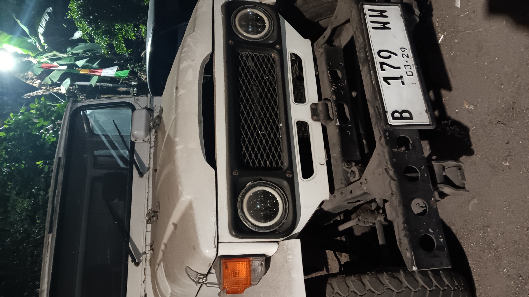 The image shows the front of a white, vintage-style SUV, likely a Toyota Land Cruiser, parked at night. The vehicle has prominent round headlights with LED rings, a black mesh grille, and an orange turn signal on the left fender. A black bumper is visible, with a license plate reading "B 179 WW" and "03-29" underneath. The license plate format suggests it is from Jakarta, Indonesia, which includes the city of South Jakarta. The vehicle is parked on a dirt or gravel surface. In the background, there are trees and foliage, illuminated by a bright light source, possibly a streetlight, which also casts shadows. There is a small Indonesian flag with red and white stripes hanging from a pole in the background, near the trees. No people are visible in the image.