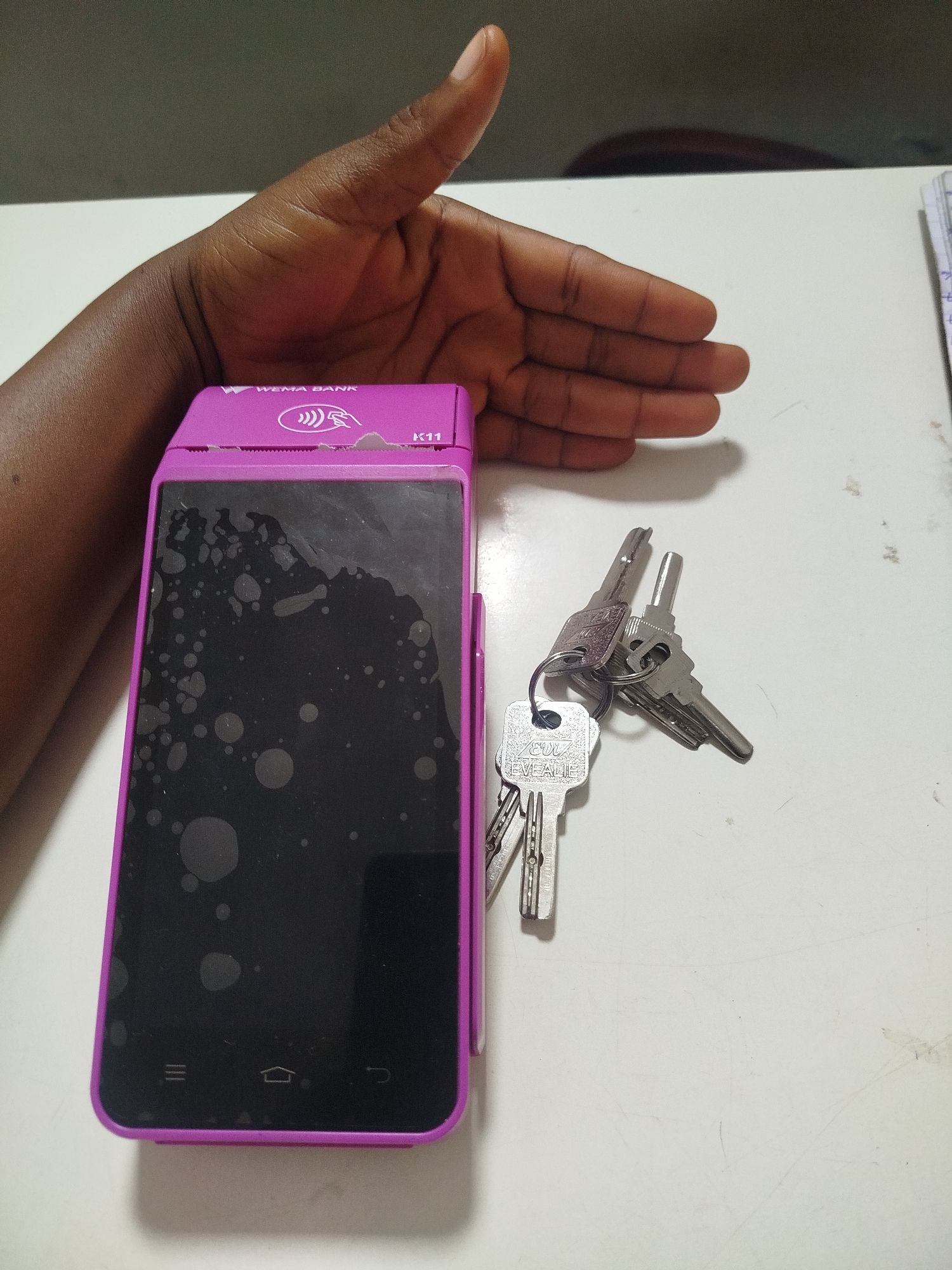 The image depicts a close-up, overhead view of a hand and a point-of-sale (POS) device on a white surface. The hand, belonging to a person with dark skin, is positioned in the upper left quadrant of the frame, palm open and facing downwards. The POS device, a purple "K11" model from "WEMA BANK," is situated in the center-left of the image, lying flat. Its screen is dark and smudged with several irregular light-colored marks, possibly water spots or fingerprints. A set of three silver keys, attached to a keyring, rests on the white surface to the right of the POS device. The keys are varied in design and appear to be house or office keys. The overall scene suggests a typical transaction or work environment. The image was taken in Uselu, Nigeria.