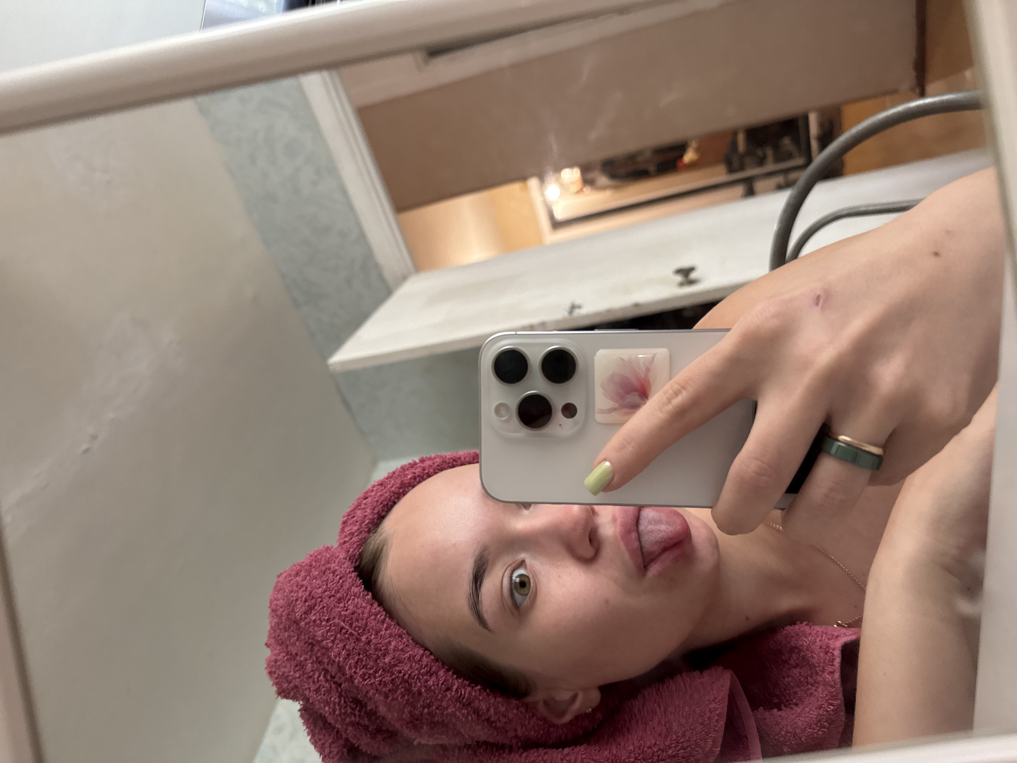 A mirrored selfie taken by a fair-skinned person in a bathroom, possibly in Chaikovsky, Russia. The person has a pink towel wrapped around their head and is sticking their tongue out. They are holding a white smartphone with three camera lenses, featuring a floral design case detail, in their right hand. A dark green ring is visible on their finger. A small scratch is visible on their right hand. The bathroom features white walls and a cabinet, and the interior of the cabinet is visible in the mirror's reflection.