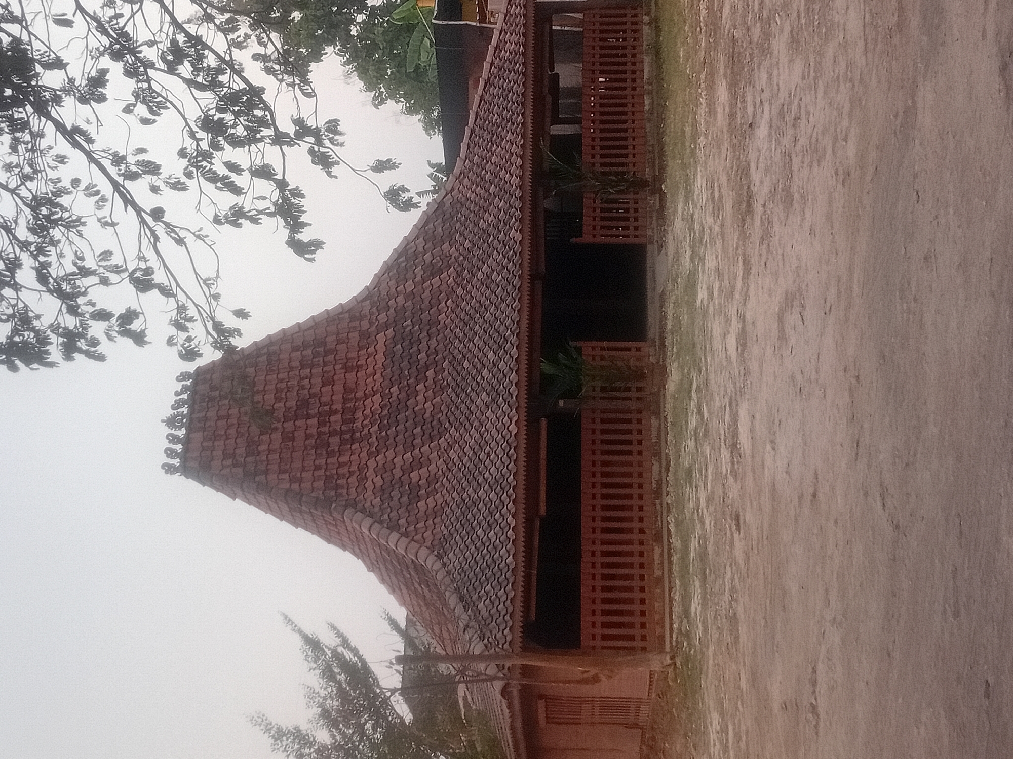The image shows a traditional Javanese house in Besito, Indonesia. The house features a distinctive tiered, curved roof covered in red tiles. A wooden fence encloses the front porch, which is open with visible dark interior spaces. The building's walls appear to be made of a reddish-brown material, possibly clay or wood. Lush green foliage and tree branches frame the top and sides of the house, suggesting an outdoor setting. The ground in front of the house is a sandy or dusty surface. The sky is overcast and hazy, contributing to a soft, diffused light. The image is captured in a static, front-facing view, with no people or active subjects visible.
