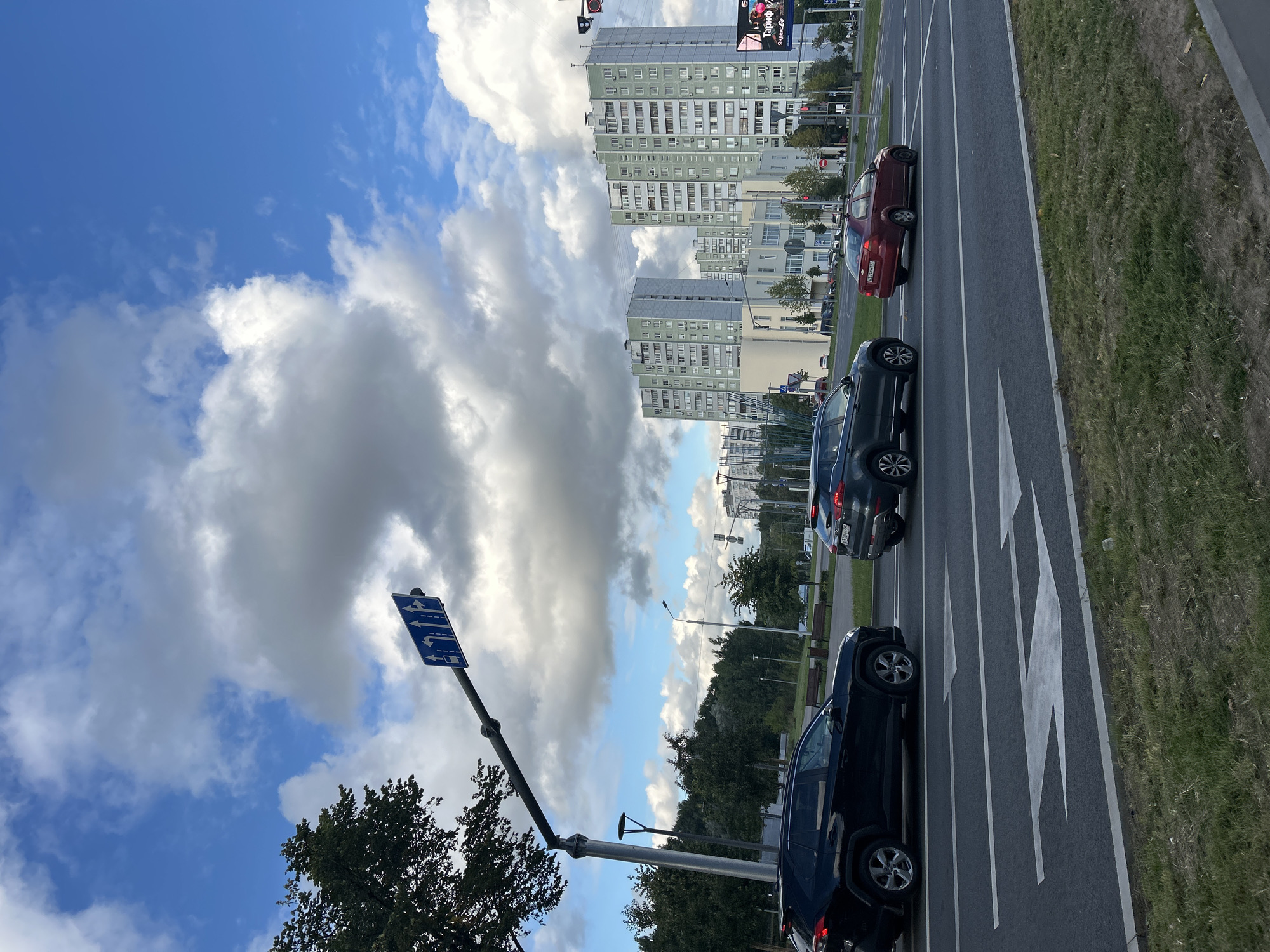 A traffic sign indicating multiple lanes and turn directions is mounted on a pole over a multi-lane road. A black SUV, a dark gray SUV, and a red sedan are driving on the road. In the background, several tall, light-green apartment buildings stand under a sky with scattered white and gray clouds. The scene appears to be in a residential area of Moscow, Russia, likely on a daytime with overcast skies.