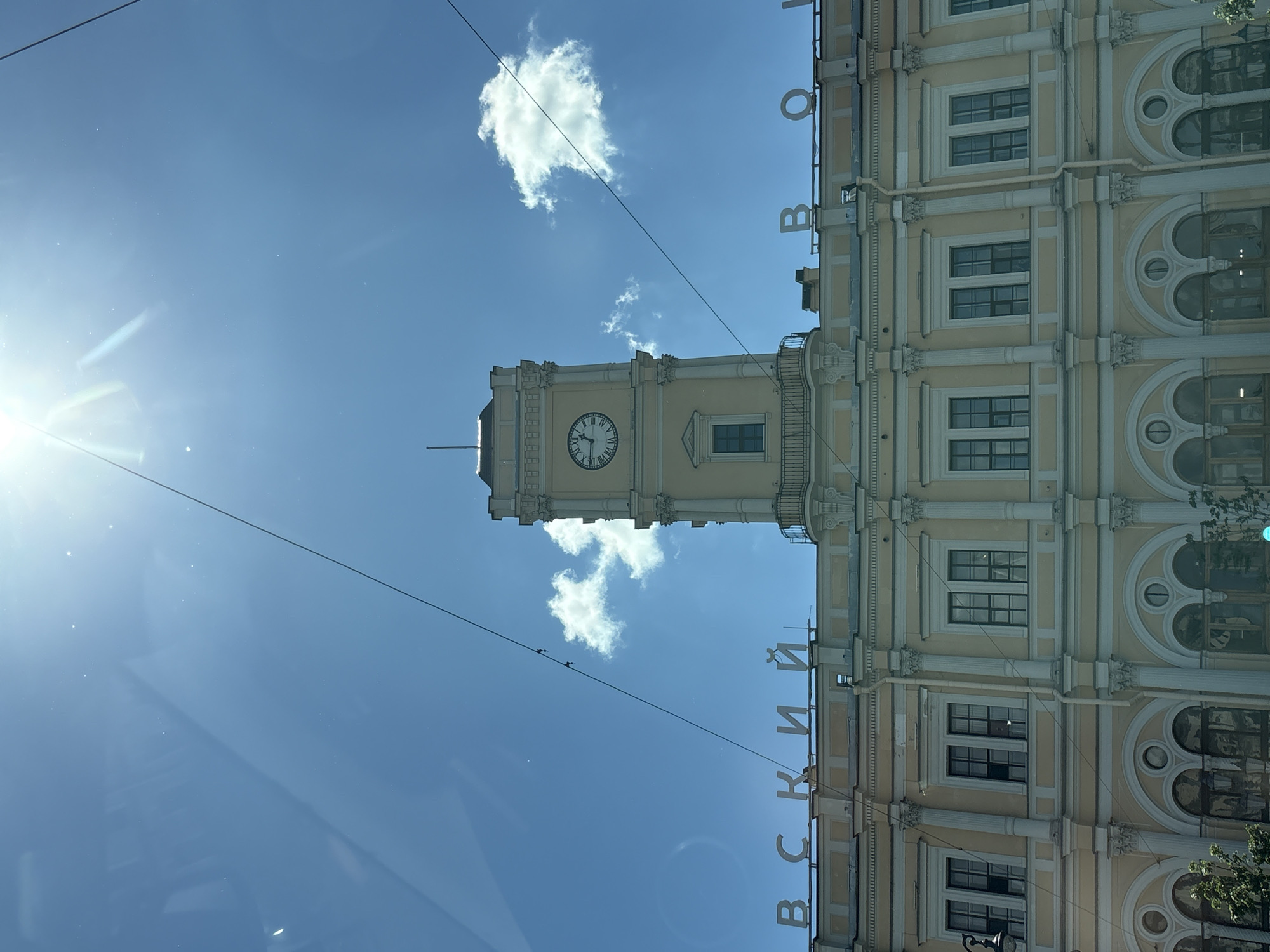This is a low-angle, full outdoor shot of the Nevsky Prospect Railway Station in Saint Petersburg, Russia, on a clear, sunny day. The architecture is neoclassical, with a pale yellow facade, white columns, and numerous arched windows. The prominent feature is a tall clock tower with a Roman numeral clock face, topped by a small spire. White letters spelling "Невский" (Nevsky) are visible across the facade on the left, and letters spelling "Вокзал" (Vokzal, meaning Railway Station) are visible on the right. The sky is a bright blue with a few wispy white clouds. The sun is high and bright, causing lens flare and glare on the image, particularly in the upper left quadrant. Electrical wires are stretched across the sky, running diagonally through the frame. Some greenery is visible at the bottom of the frame.