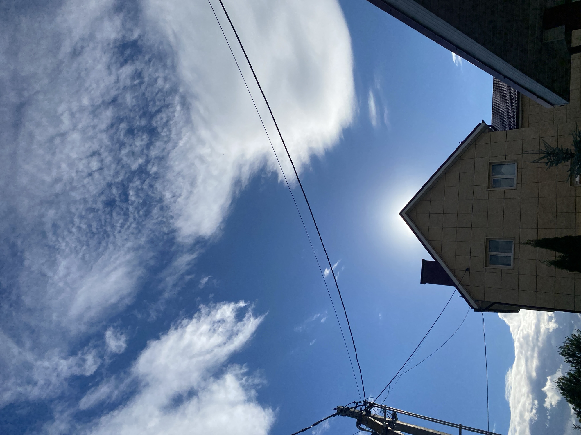 The image depicts a bright, sunny day with a clear blue sky dominating the upper portion. Wispy, cirrus clouds are scattered across the sky, with a larger, more defined cloud formation on the right. The sun is directly behind the peak of a two-story house, creating a silhouette effect and a sunburst that obscures the exact details of the roofline and chimney. The house appears to be clad in light-colored square tiles. Two windows are visible on the upper floor of the house, reflecting the bright sky. To the left of the house, a utility pole with wires is visible. In the foreground, on the right side of the image, part of another building's roof is visible, casting a shadow. Two small, dark green evergreen trees are planted in front of the house. The scene appears to be a residential area.