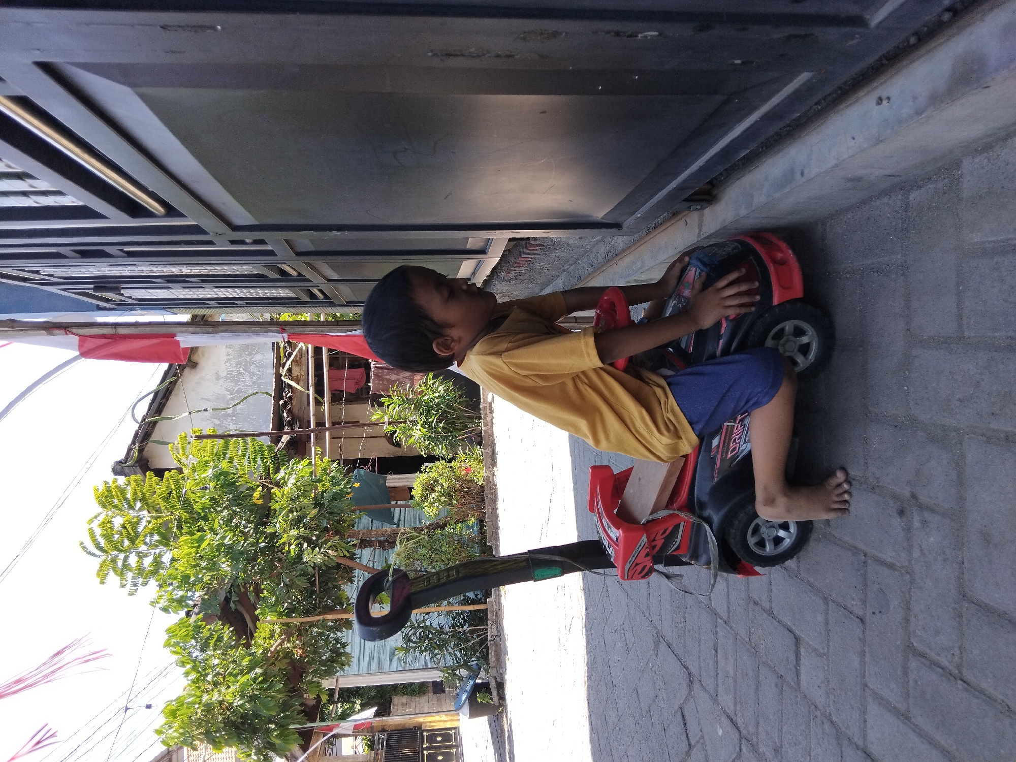 A young boy wearing a yellow t-shirt and blue shorts is positioned on a red and black toy car on a paved walkway. The boy is facing towards a dark metal gate, with his right knee bent and his left leg extended. His hands are resting on the front of the toy car. The toy car has a handle at the back. Behind the boy and to the left, there are trees with green leaves, some houses with tiled roofs, and flags. The ground is made of interlocking gray bricks. The scene appears to be outdoors in a residential area, likely in Kepuh Kiriman, Indonesia.