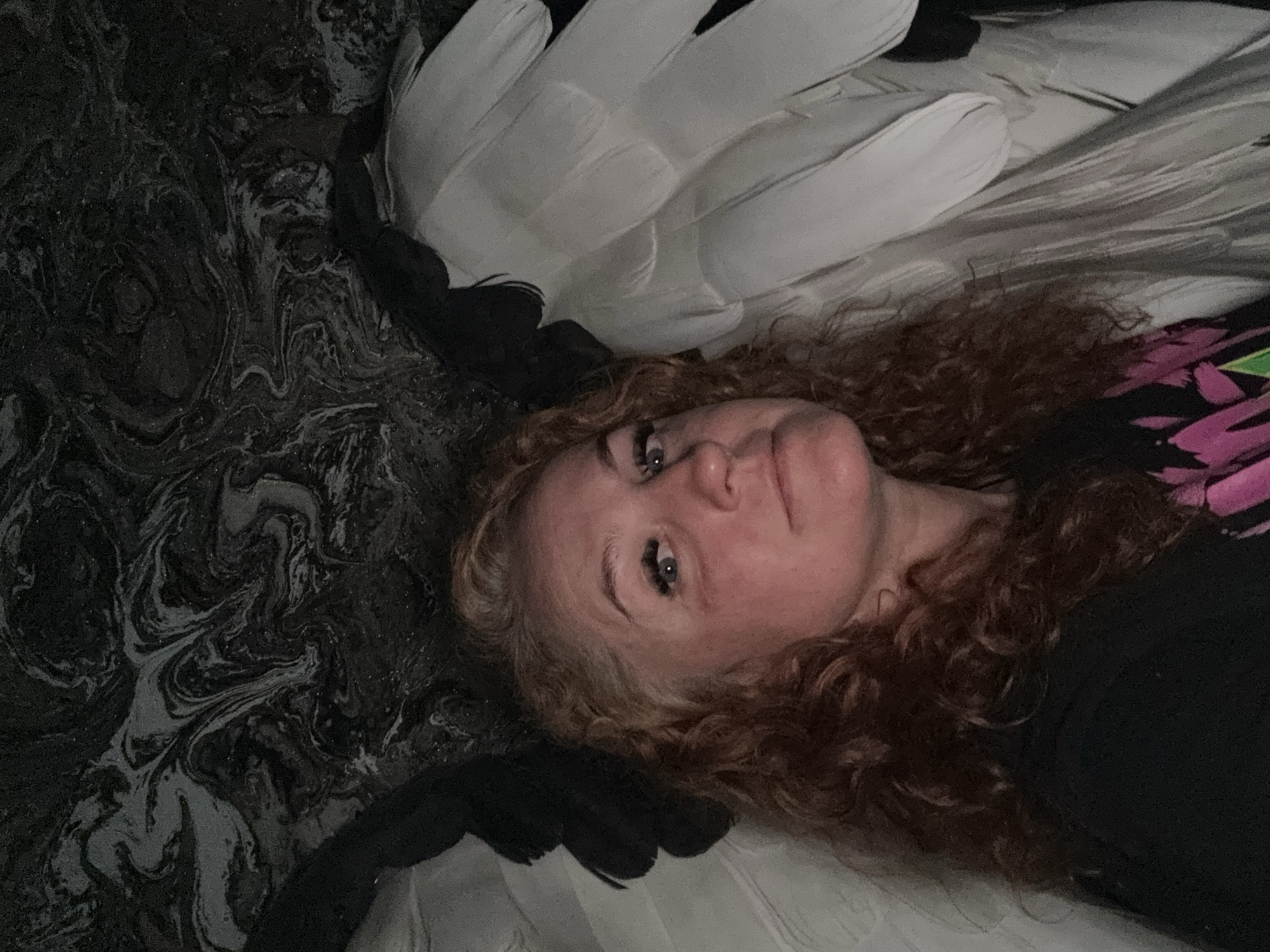 A woman with light skin and curly, reddish-brown hair is lying on her back. She has blue eyes and wears false eyelashes. Above her head are large, white feathers with black tips, possibly part of a costume or decorative piece. The background consists of two distinct patterns: a swirling, marbled black and gray design on the left and a striped white and gray pattern on the right. The woman wears a dark-colored shirt with a pink and green design visible on the right side of the frame. The scene appears to be indoors. There is no verifiable information about the location being in the city of Chaikovsky, Russia.