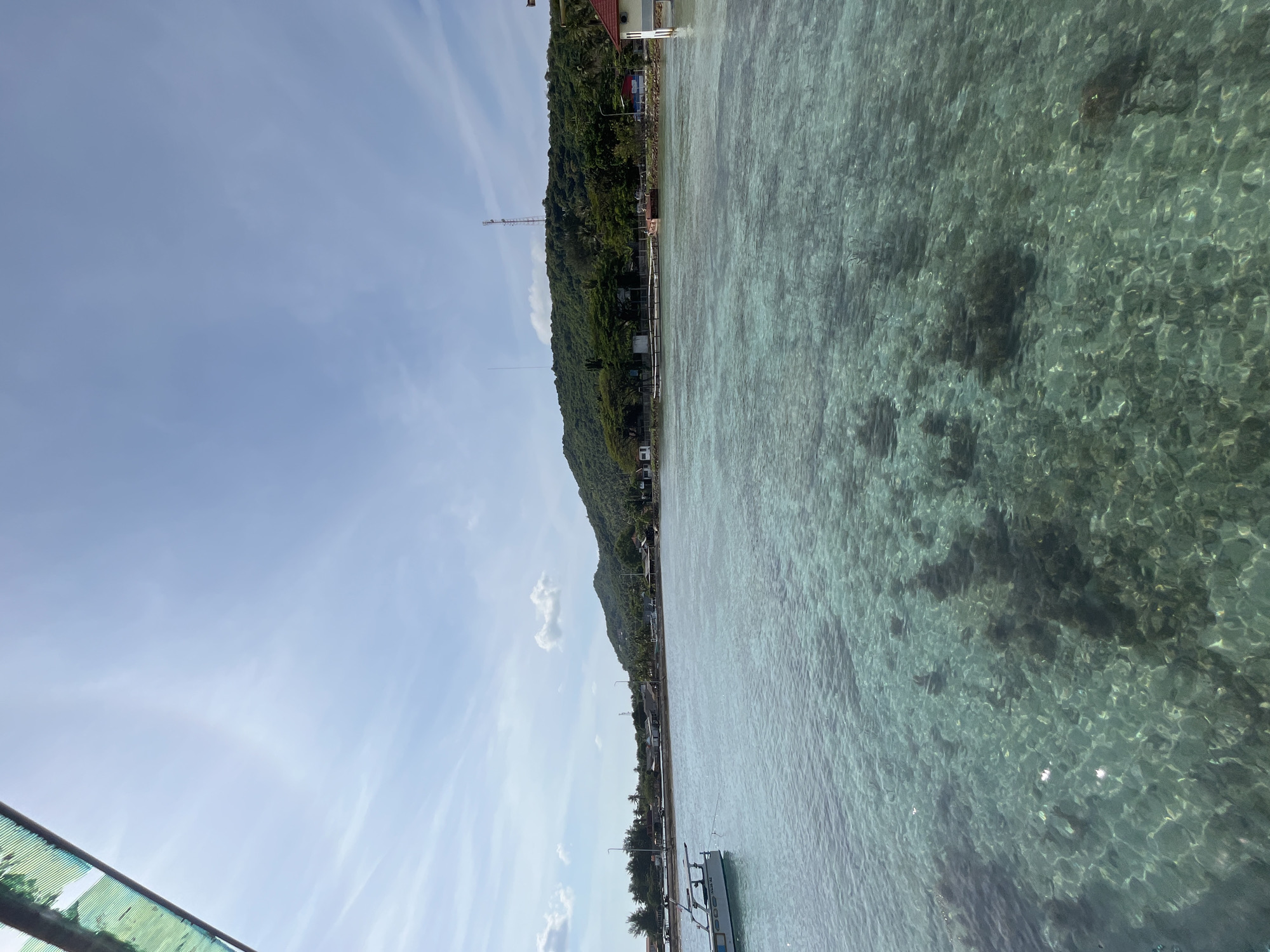This is a scenic photograph of the coast of Karimunjawa, Indonesia, showcasing the clear, shallow turquoise waters with visible coral formations beneath the surface. The foreground is dominated by the translucent sea, reflecting the sunlight and revealing the textured seabed. In the middle ground, a small white boat is moored, its hull partially submerged. Beyond the water, a low-lying coastline is visible, characterized by a strip of buildings and vegetation. A treeline separates the structures from a lush, green, hilly landscape that forms the backdrop of the scene. The sky is a bright, pale blue with scattered wispy clouds and a faint rainbow arc visible in the upper left quadrant. The overall impression is one of a serene tropical coastal environment.