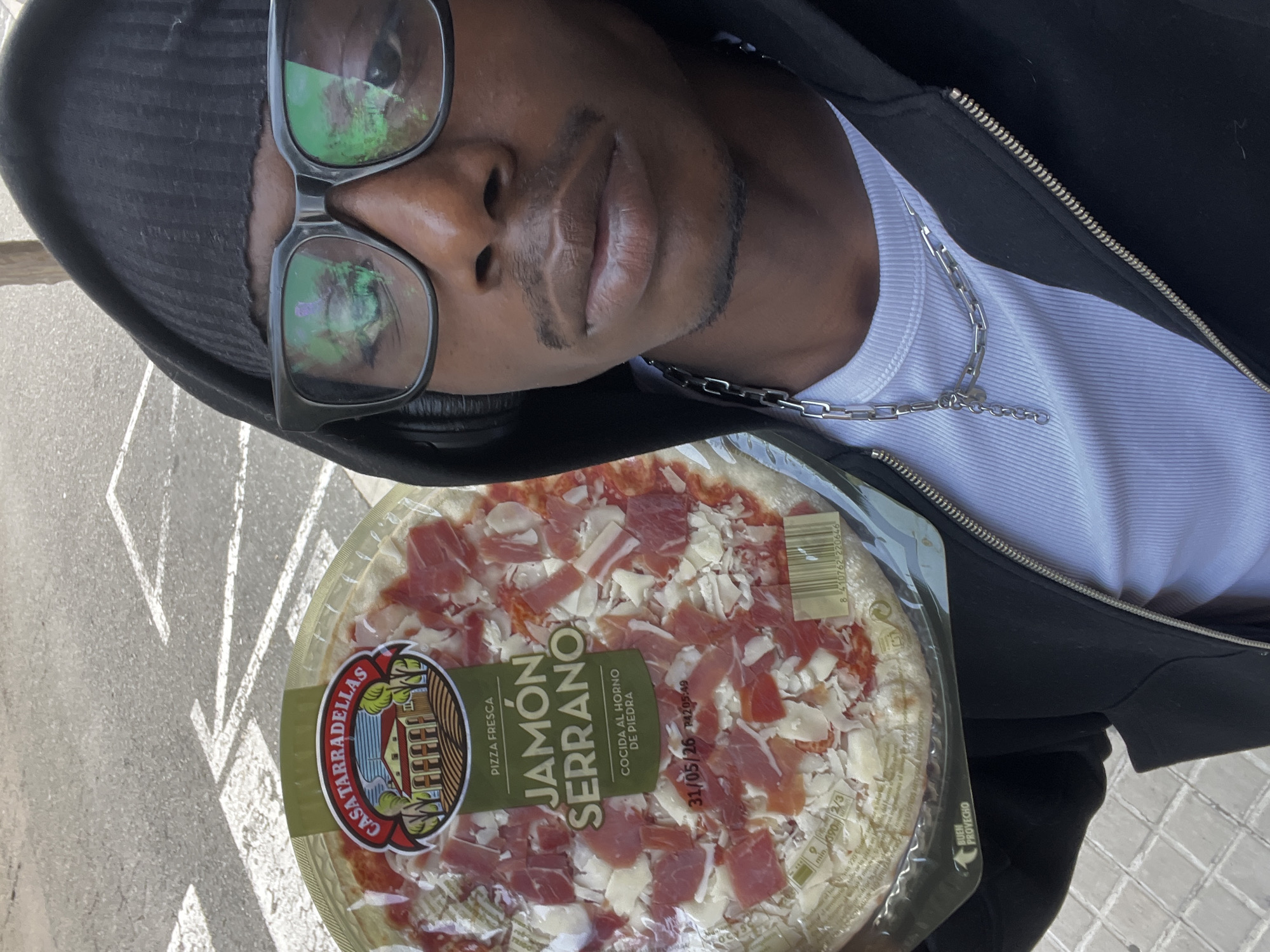 An individual with dark skin wearing a black hooded jacket, a white ribbed tank top, and black-framed glasses is holding a packaged "Casa Tarradellas Jamon Serrano" pizza. The pizza is topped with jamón serrano, cheese, and a red sauce. The labels on the pizza are in Spanish. The individual is wearing a silver chain necklace. The setting appears to be an outdoor area in Barcelona, Spain, based on the style of the pizza. The background includes a light-colored pavement.