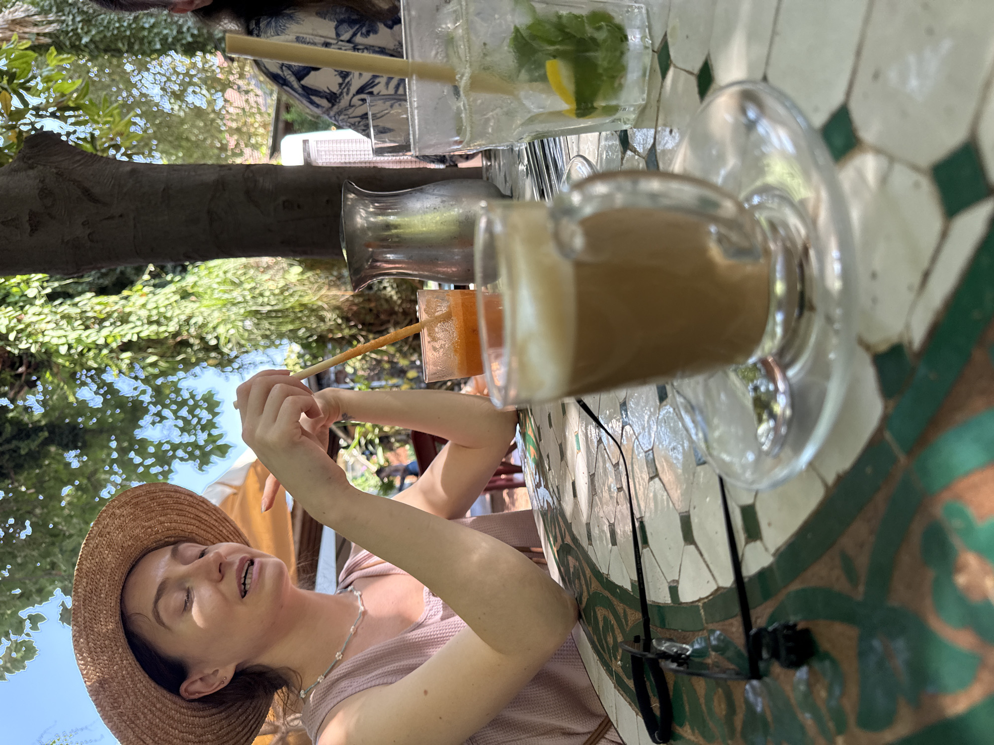 A woman wearing a straw hat and a pink top is sitting at an outdoor table. She is looking to her right with her eyes closed and her mouth slightly open, as if speaking or taking a sip. She is holding a straw in her right hand, which is in a glass of orange juice. In front of her is a glass of coffee with foam. On the table are other drinks, a metal pitcher, and a pair of glasses. The table has a patterned surface with green and white tiles. The background shows greenery and trees, suggesting an outdoor setting in a location like Mevoot HaHermon Regional Council, Israel.