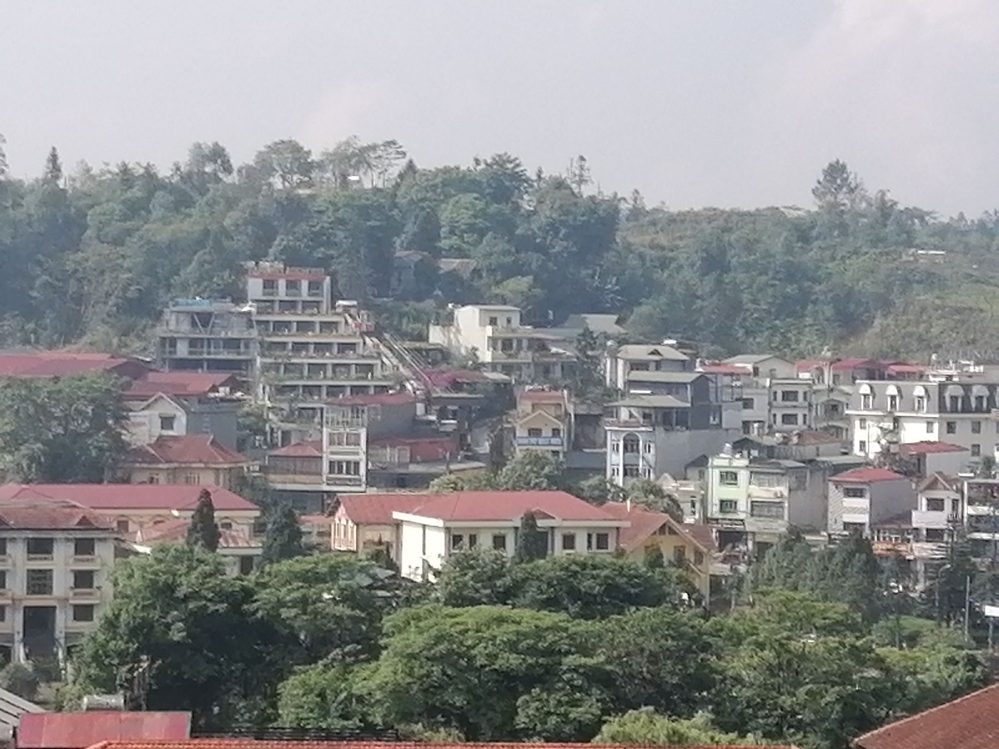 A daytime, high-angle shot captures a portion of Sa Pa Ward, Vietnam. The foreground features dense, green trees interspersed with buildings that have red-tiled roofs. Beyond this, multiple buildings of varying heights and architectural styles occupy the midground, exhibiting a mix of white, beige, and blue facades. Some structures appear to be under construction or in a state of disrepair. The background consists of a hillside densely covered in green foliage, partially obscured by haze. The sky is overcast. No people are visible.