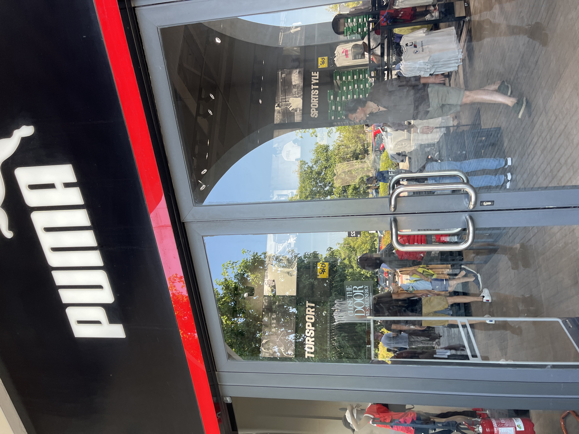 The image shows the exterior of a Puma store in Rodengo, Italy. The store has a black facade with the white Puma logo and name prominently displayed above the entrance. The entrance consists of large glass doors with silver handles. Reflected in the glass doors are trees, other buildings, and pedestrians. Inside the store, racks of clothing and shoe boxes are visible. A fire extinguisher is mounted to the left of the entrance. The overall scene depicts a typical retail environment on a sunny day.