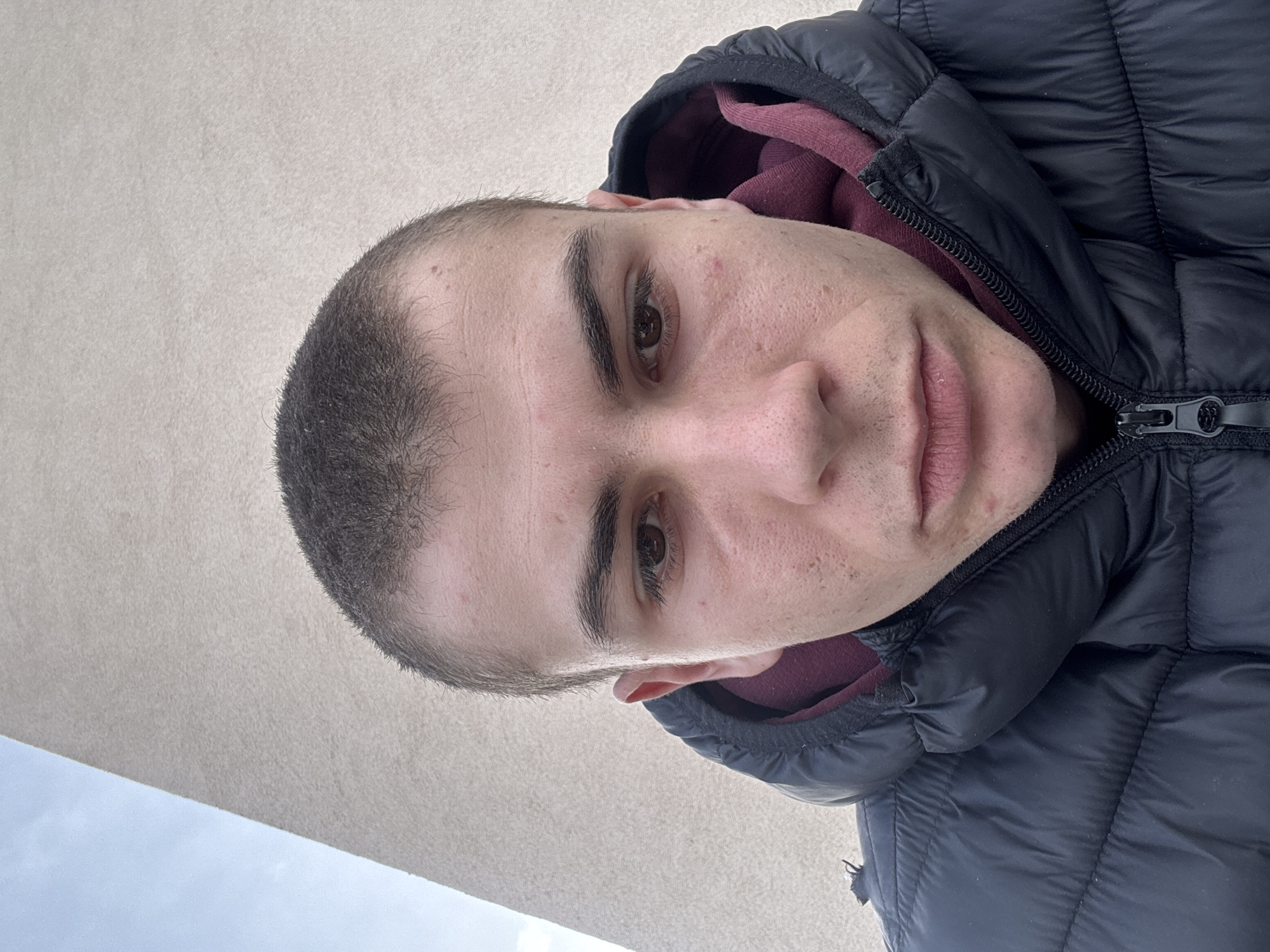 A close-up, angled portrait shows a light-skinned male wearing a puffy, black jacket over a maroon hooded sweatshirt. The man has short, dark hair and brown eyes. His face shows minor skin imperfections. The background includes a cream-colored wall and a glimpse of a light blue sky. No specific details indicate the location is Plovdiv, Bulgaria; this information is not verifiable from the image.