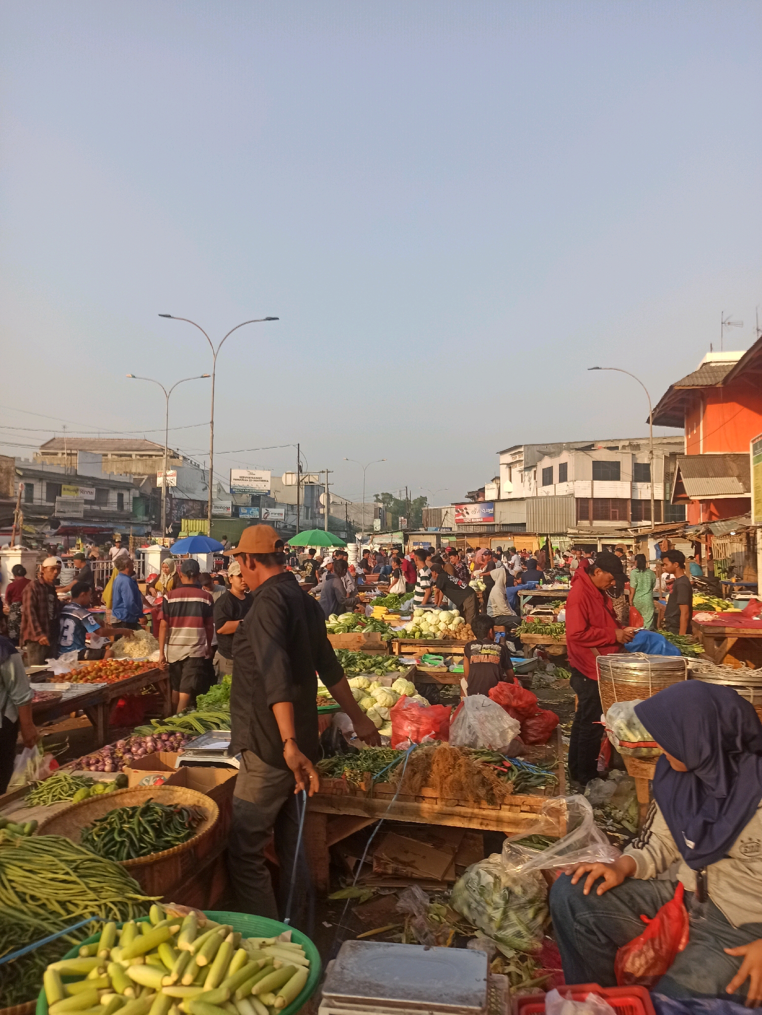 This is a daytime, medium-shot, eye-level photograph of a busy outdoor market scene in Cikarang, Indonesia. The sky is clear and pale blue, with streetlights visible in the background. The foreground and mid-ground are filled with numerous people, predominantly adults, engaged in various activities related to commerce.
The market appears to be selling fresh produce, with stalls laden with vegetables such as long beans, corn, cabbage, onions, and peppers. Wicker baskets and plastic bags are used to hold and display the goods. Several wooden tables and makeshift stalls are set up to organize the produce. People are seen interacting with each other, presumably as vendors and customers. Some individuals are wearing traditional Indonesian attire, including headscarves.
In the mid-ground, several buildings line the street, some of which appear to be shops or businesses with signage. The architecture suggests a semi-urban environment. The ground is paved, and debris and discarded packaging are visible in some areas, indicating a typical bustling market atmosphere. The overall impression is one of a lively and active marketplace.