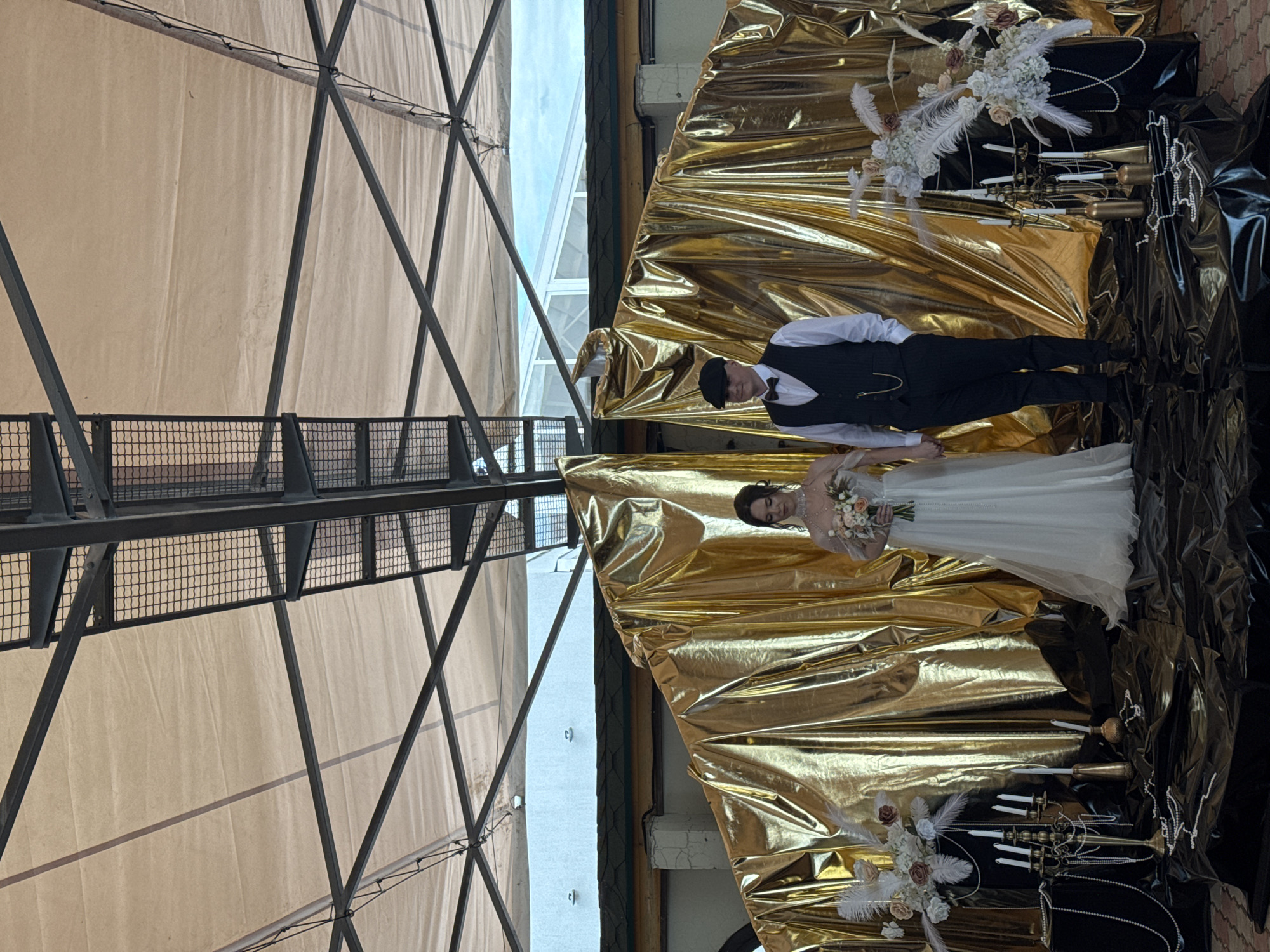 A forensic image depicts a full-shot of a couple, identified as a bride and groom, standing centrally on a decorated stage under a large fabric canopy supported by a dark metal truss structure. The location is in Unknown, Unknown. The backdrop behind the couple is a large, golden, draped fabric, appearing to be two panels slightly parted in the middle. The ground directly beneath the couple and surrounding decorations is covered with crumpled black, shiny fabric.
The bride, positioned on the left, is wearing a long-sleeved white wedding gown with a sheer upper bodice and a full skirt. She has dark hair styled up and wears a pearl choker necklace. She holds a bouquet of light-colored flowers with peach and pink accents in her left hand. Her right hand is clasped with the groom's left hand.
The groom, on the right, wears a white collared shirt, a black vest, a black bow tie, dark trousers, and a black flat cap. A watch chain is visible extending from his vest pocket. His right hand is in his trouser pocket.
Flanking the couple on the black draped floor are elaborate decorations. On both the left and right, tall dark pedestals support large floral arrangements featuring white and light-colored flowers (roses, hydrangeas), interspersed with prominent white feathers and subtle reddish-brown foliage. Multiple strands of white pearls are draped around these pedestals and across the black fabric. Several white taper candles are arranged in ornate golden or bronzed candelabras and individual holders. Small golden bottles or vases are also present.
Above the golden backdrop, sections of a light-colored building facade are visible on the left, while a blue sky with clouds and parts of a white/glass structure are seen on the right, suggesting an outdoor or semi-outdoor venue. The overhead canopy is a light, possibly beige, fabric, stretched taut and supported by the dark metal framework, some parts of which include wire mesh. The overall lighting is bright and even.