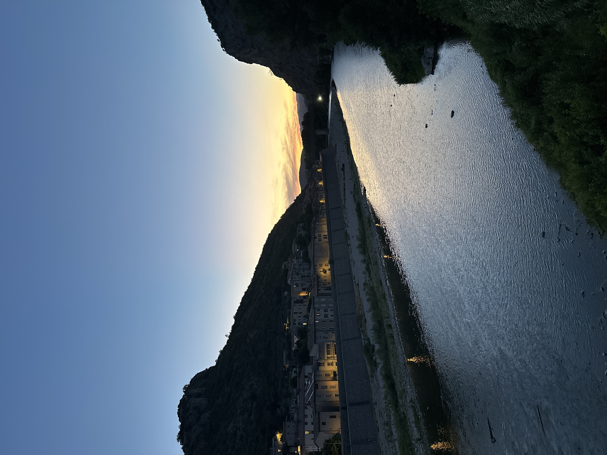 The image depicts a scenic view of the Gardon River in Anduze, France, at dusk. A row of illuminated buildings lines the riverbank, nestled against a dark, tree-covered hillside on the left. On the right, a steep, rocky cliff face rises from the water's edge, also covered in dense vegetation. The river itself flows gently, reflecting the twilight sky and the distant lights from the town. The sky is a clear, gradient blue, transitioning to a warm yellow and orange band near the horizon where the sun has recently set. A bridge can be seen in the distance crossing the river. There are no people visible in the image. The overall atmosphere is tranquil and picturesque, showcasing the natural beauty of the area alongside the charming architecture of the town.