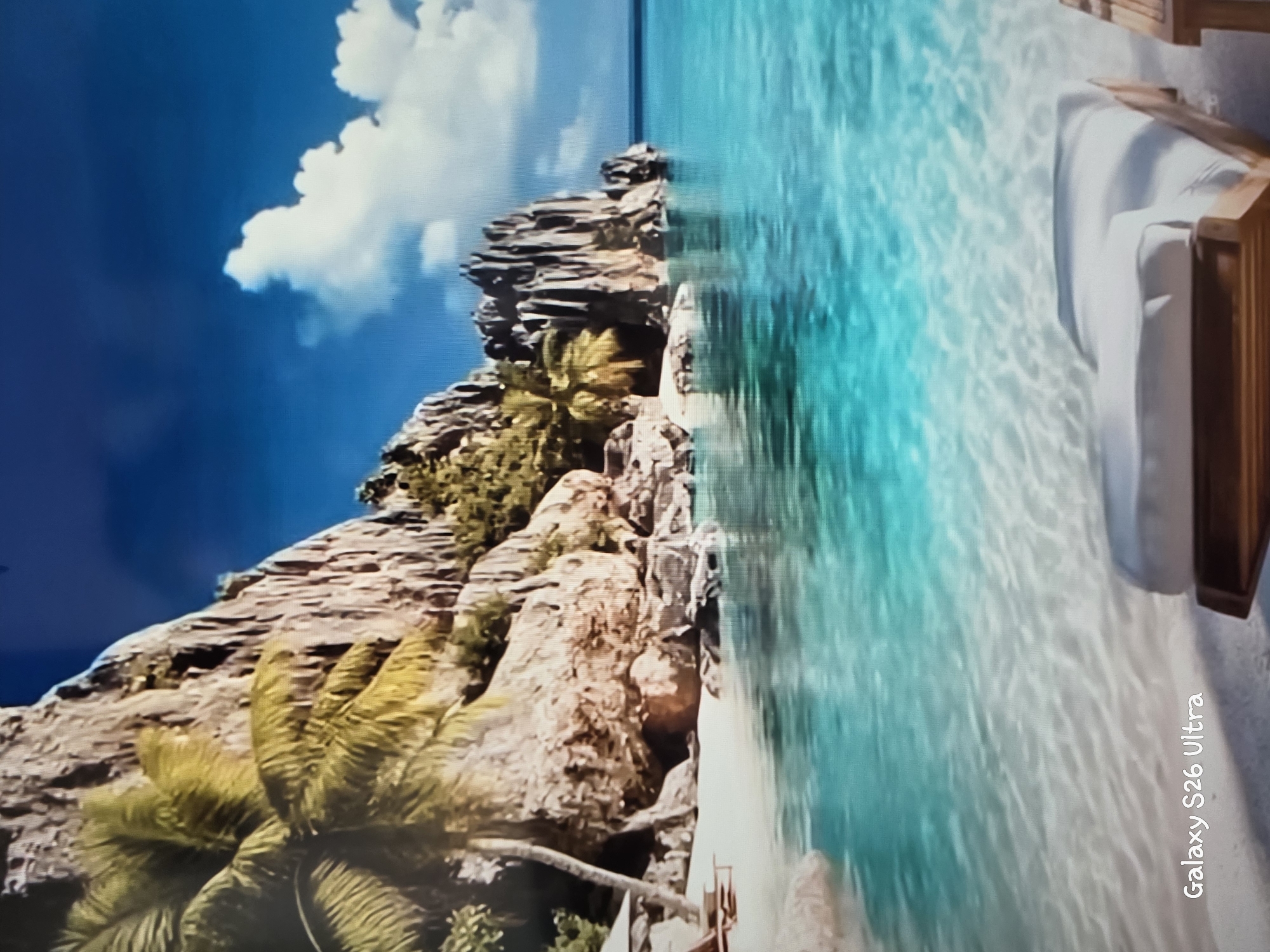 This is a photograph of a tropical beach scene. In the foreground, there is clear turquoise water with ripples on the surface, revealing the sandy bottom. A portion of a white lounge chair with a wooden frame is visible on the sand at the lower right. In the middle ground, a white sand beach meets the water, with several lounge chairs and umbrellas. Behind the beach, a rocky cliff face rises, adorned with palm trees and other tropical vegetation. The sky above is a bright blue with scattered white clouds. The image contains a watermark in the lower left corner that reads "Galaxy S26 Ultra". This image does not depict any location in the city of Plovdiv, Bulgaria.