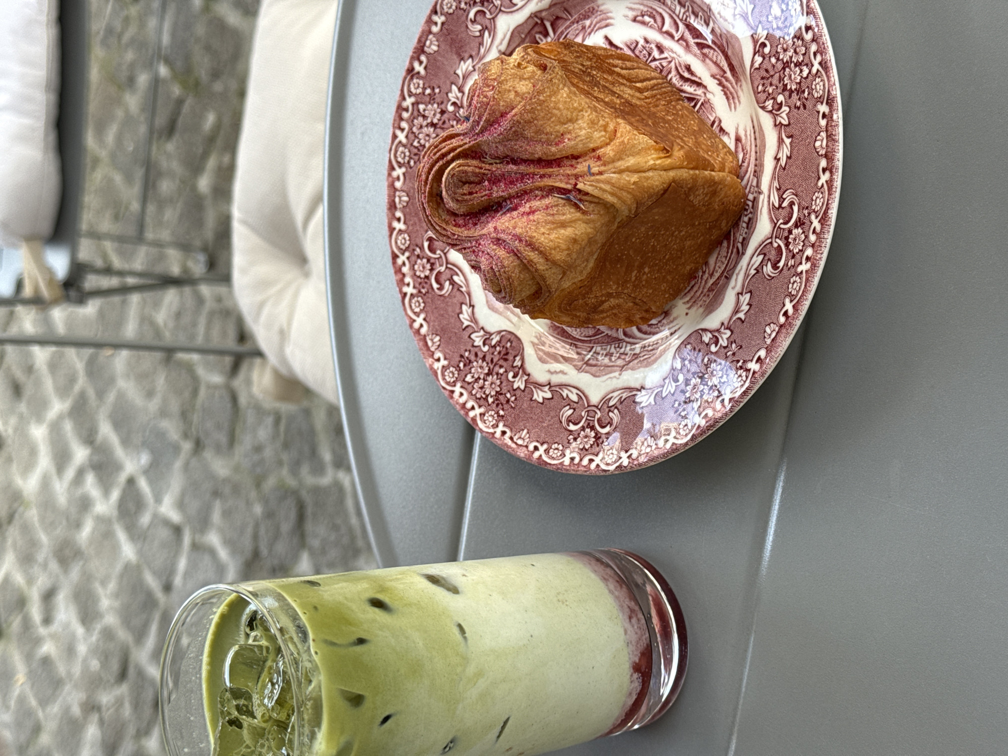 An overhead shot shows a glass of iced matcha latte with a layer of berry puree at the bottom, positioned on a grey table next to a decorative plate containing a golden-brown pastry with pink topping. The setting appears to be an outdoor cafe in Vracar Urban Municipality, Serbia, with cobblestone pavement and outdoor seating visible in the blurred background.