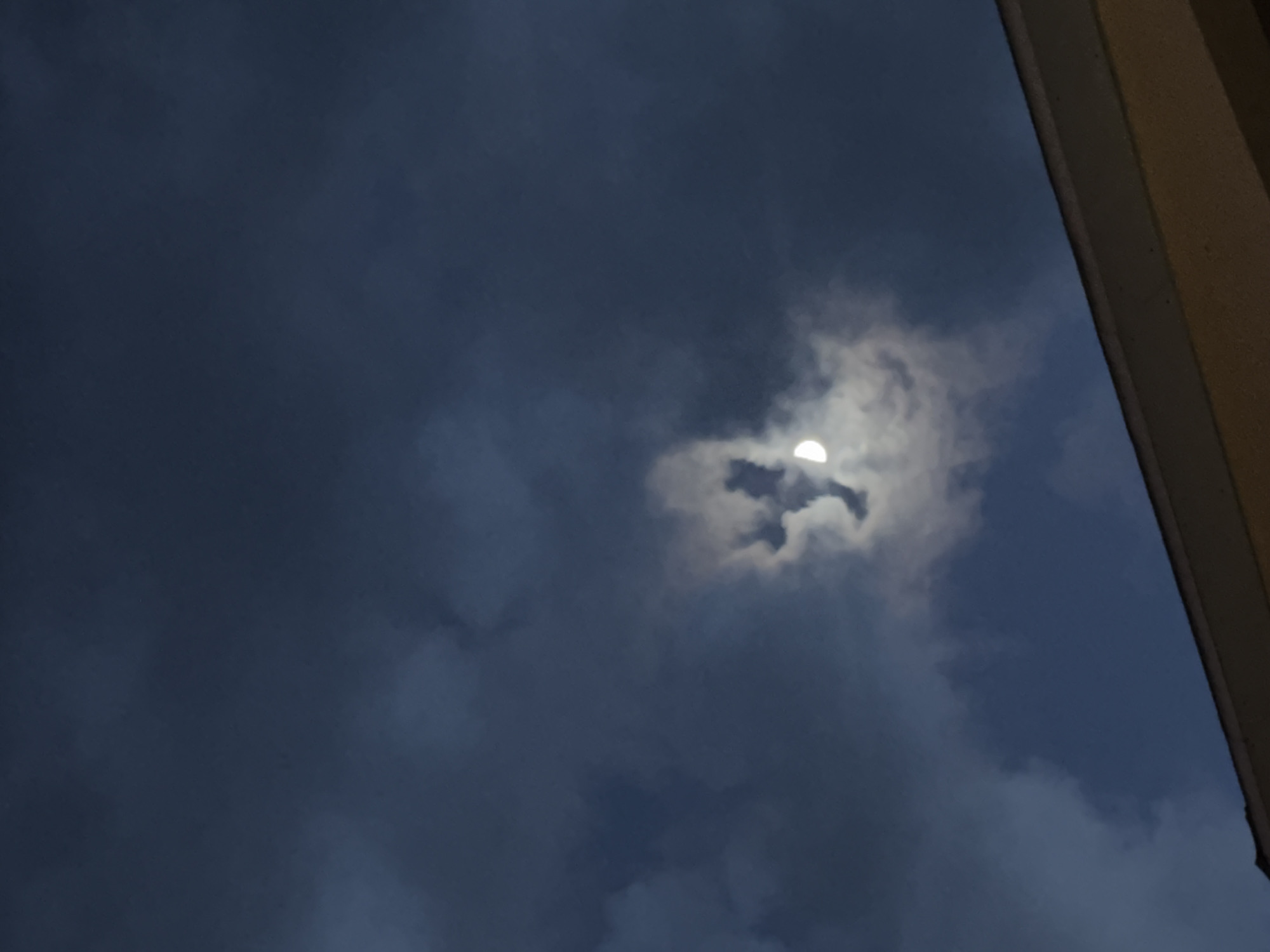 The image depicts a twilight sky with dark, heavy clouds covering much of the view. A half-moon is visible, partially obscured by a patch of brighter, white clouds that create a halo-like effect around it. The sky ranges from a deep blue-gray to lighter shades where the clouds are thinner. A portion of a building, likely a wall or pillar, is visible in the upper right corner, colored beige or light brown. The location is Unknown, Unknown.