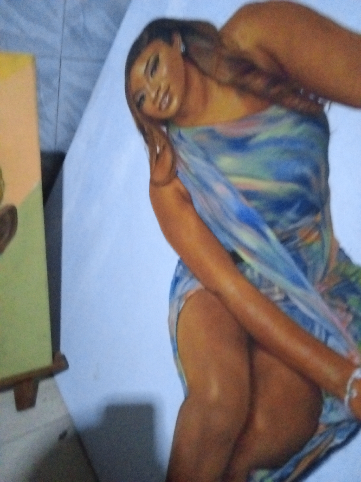 The image shows a painting of a woman. The woman in the painting is wearing a blue and green dress. A painting is visible on the left with a green and yellow background. The background appears to be a wall with a blue-ish color. The location is Ado, Nigeria.