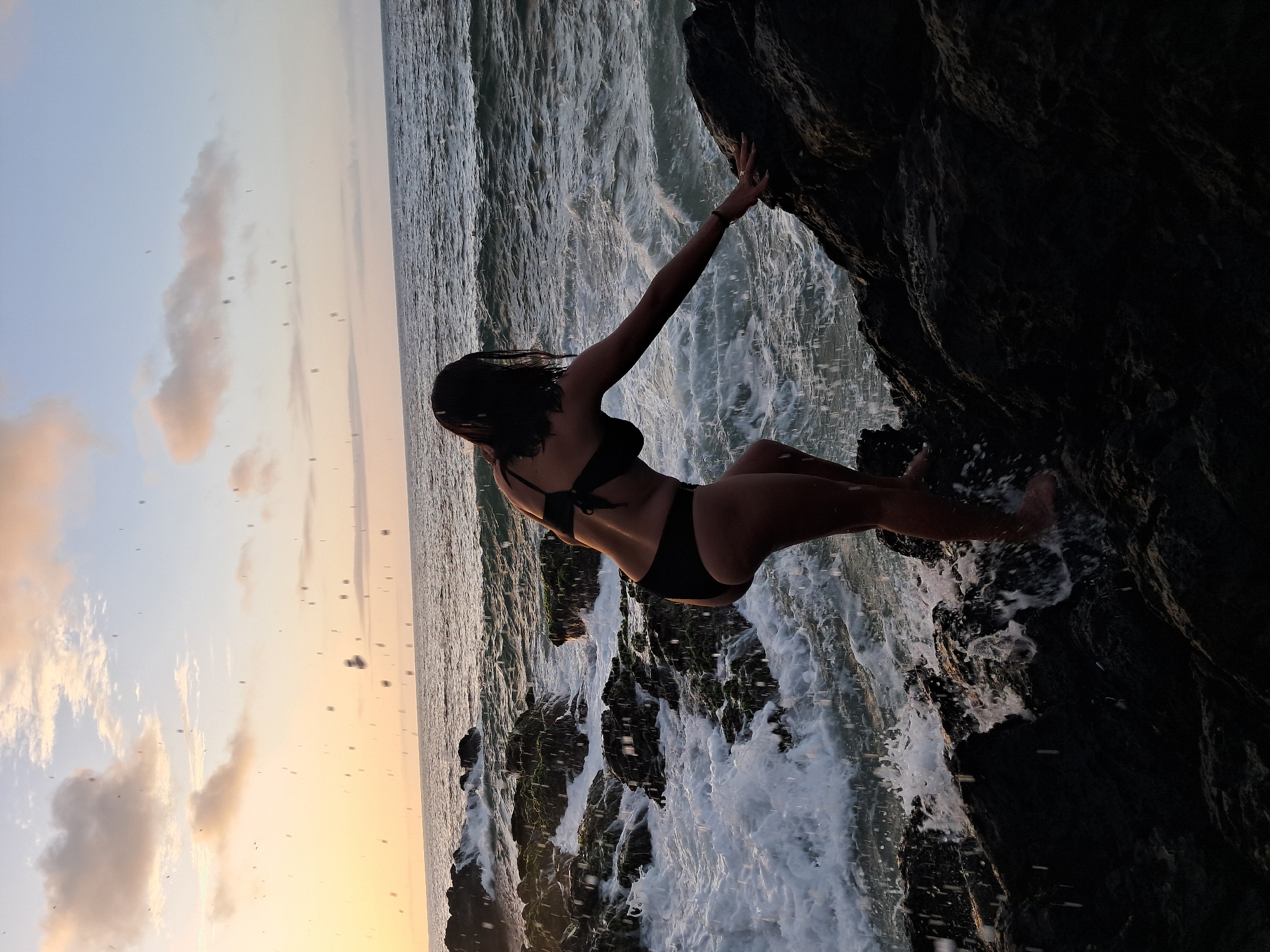 A woman in a black bikini is climbing on dark, wet rocks near the ocean at sunset in Pedro González, Venezuela. The sky is a soft gradient of orange and blue, with scattered clouds and what appear to be water droplets suspended in the air. Waves are crashing against the rocks, creating white foam. The woman is facing away from the camera, with her right arm extended to steady herself on a rock. Her hair is dark and appears wet. The overall scene is one of dynamic natural beauty and a moment of active engagement with the environment.
