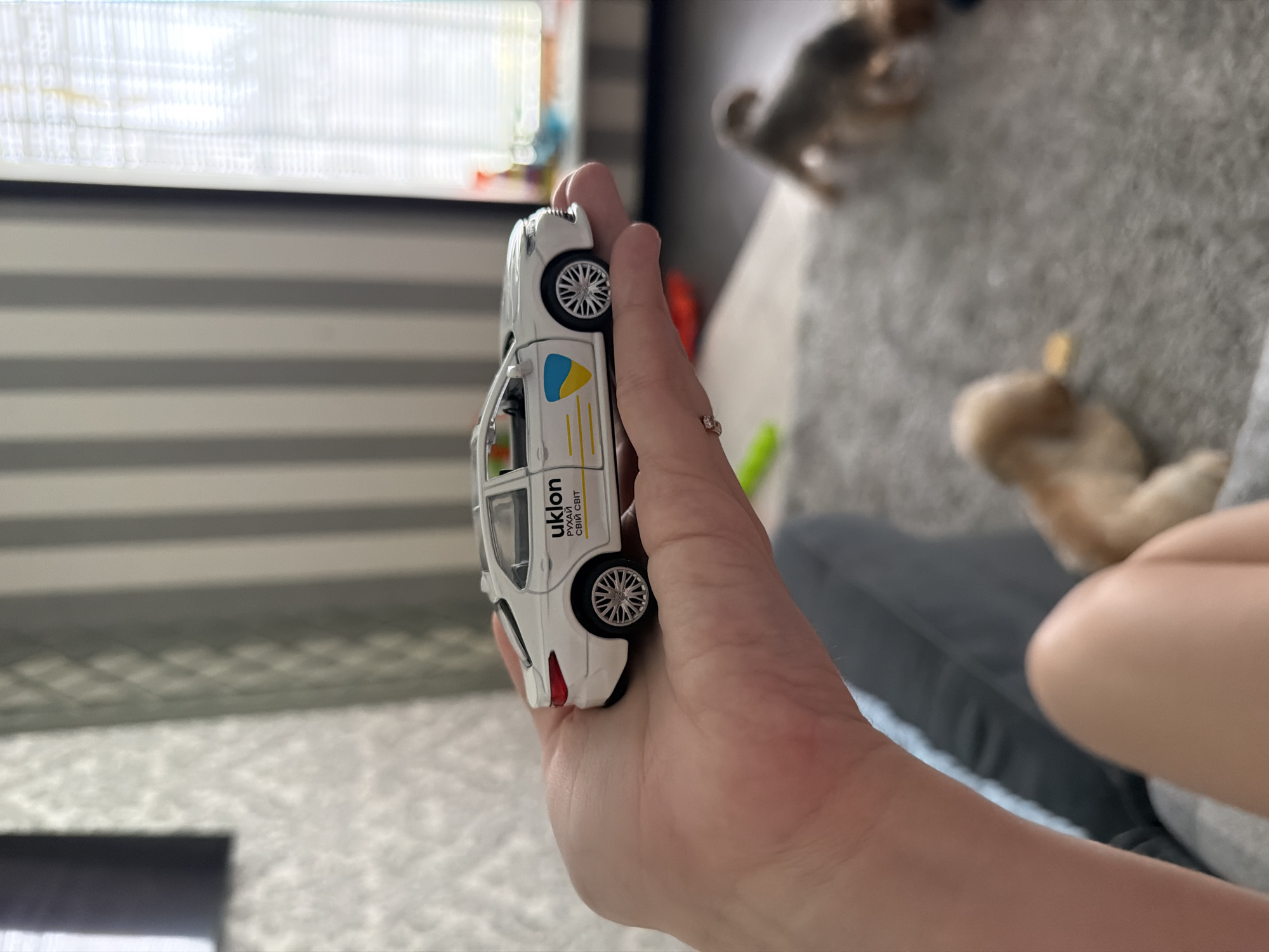 A close-up, side-profile shot shows a white toy car resting on a person's palm. The car features the "uklon" logo, a Ukrainian flag emblem, and yellow racing stripes. In the background, a blurred scene reveals two dogs on a grey carpet, and a striped wall with a window. The overall environment suggests an indoor setting within a city.