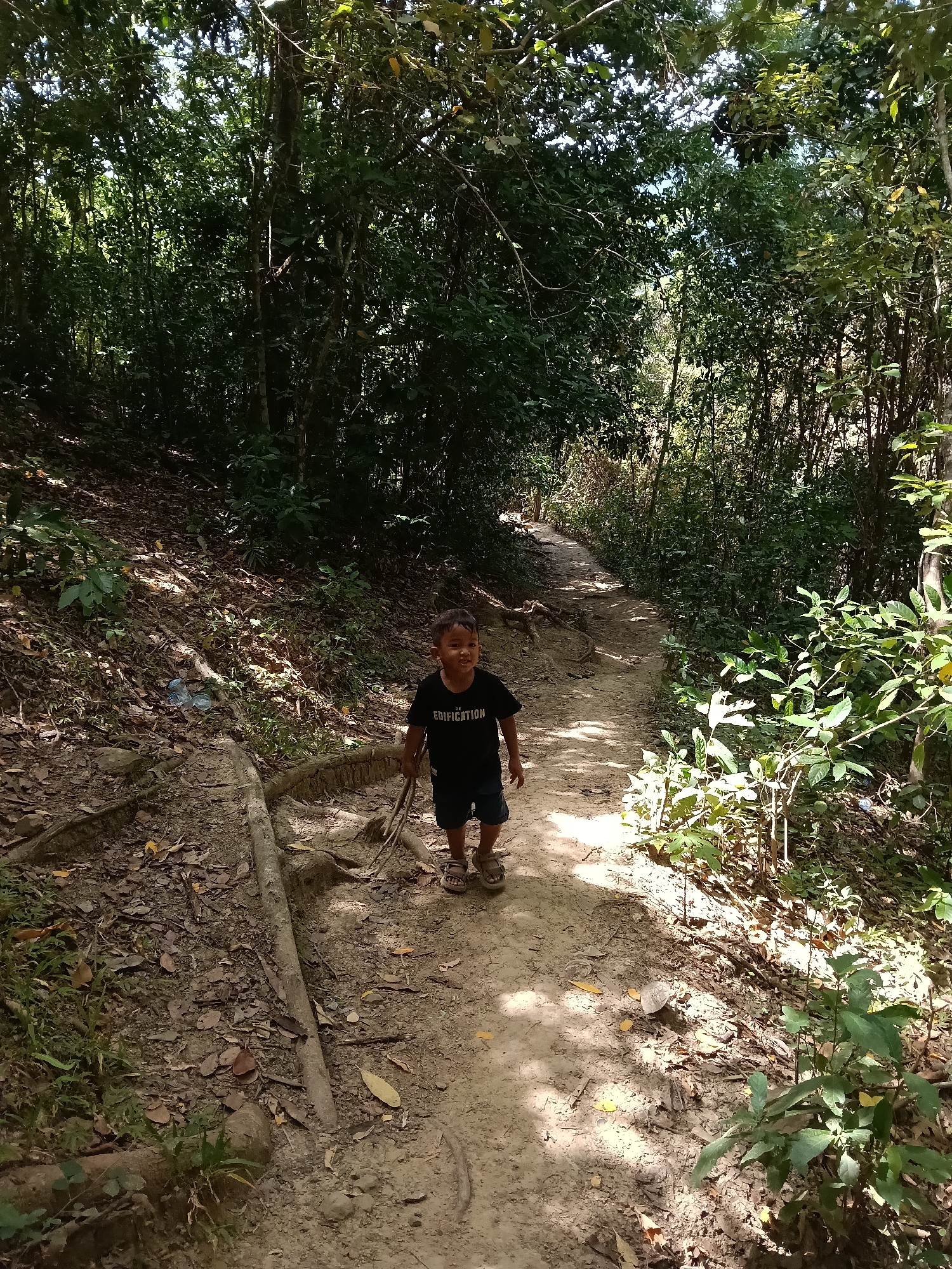 A young boy in a black t-shirt with "EDIFICATION" printed on it, and dark shorts, walks along a dirt path in a wooded area. He is smiling and looking towards the camera. The path is winding and surrounded by lush green foliage, trees, and exposed roots. Sunlight filters through the canopy, casting dappled shadows on the ground. The overall scene suggests a casual walk in nature. The location is identified as Lampageu, Indonesia.