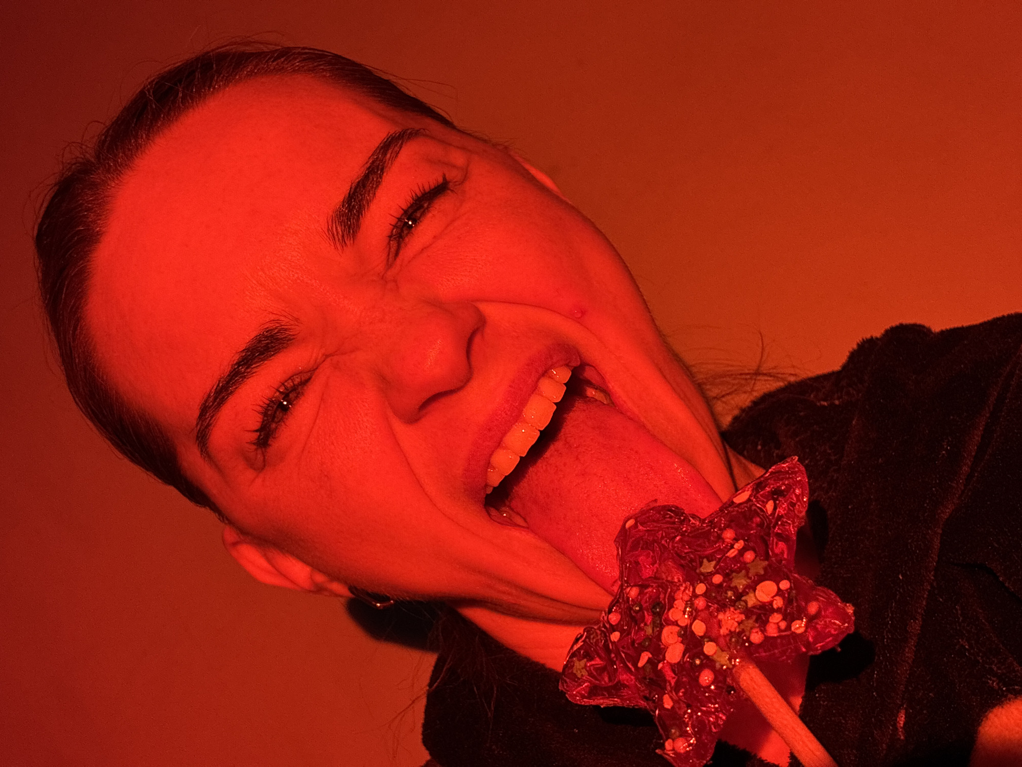 A person, likely female, is photographed under red lighting, with her head tilted and tongue extended, holding a star-shaped lollipop. Her eyes are squinting, suggesting a grimace or playful expression. The background is plain, and the subject is partially wearing a dark-colored garment. Location is not discernible from the image.