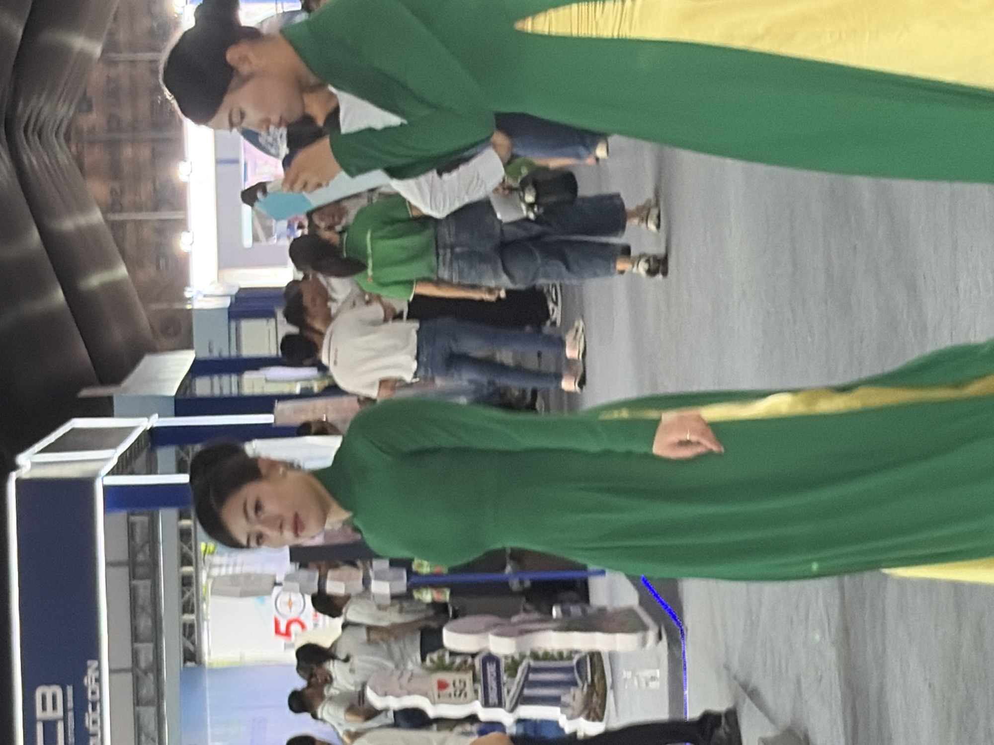 The scene depicts an indoor event or exhibition within Thủ Đức City, Vietnam. In the foreground, two women in traditional Vietnamese green Ao Dai dresses with yellow lining are visible. The woman on the left stands facing slightly to her right, her expression neutral. The woman on the right is positioned in profile, looking left, and holding a light blue rectangular object, possibly a mobile phone or tablet. Behind them, numerous people are scattered throughout the hall, some wearing white t-shirts, others green t-shirts, and casual attire, suggesting a mix of staff and attendees.
Prominent objects in the midground include display stands and signage. A white cutout display on the left features a red heart icon followed by "SG" (likely representing Singapore) and the word "SINGAPORE" below it. Another sign displays the number "50". Further back, a large blue banner with the partial text "CB" and "ƯỚC DÂN" (suggesting Vietcombank) is mounted on a structure. The environment features a grey floor, a blue guiding line, and overhead industrial-style roofing with exposed trusses and lighting fixtures, characteristic of a large convention or exhibition center. A woman in the midground wears a green t-shirt, blue wide-leg jeans, and carries a black bag, walking away from the viewer.
